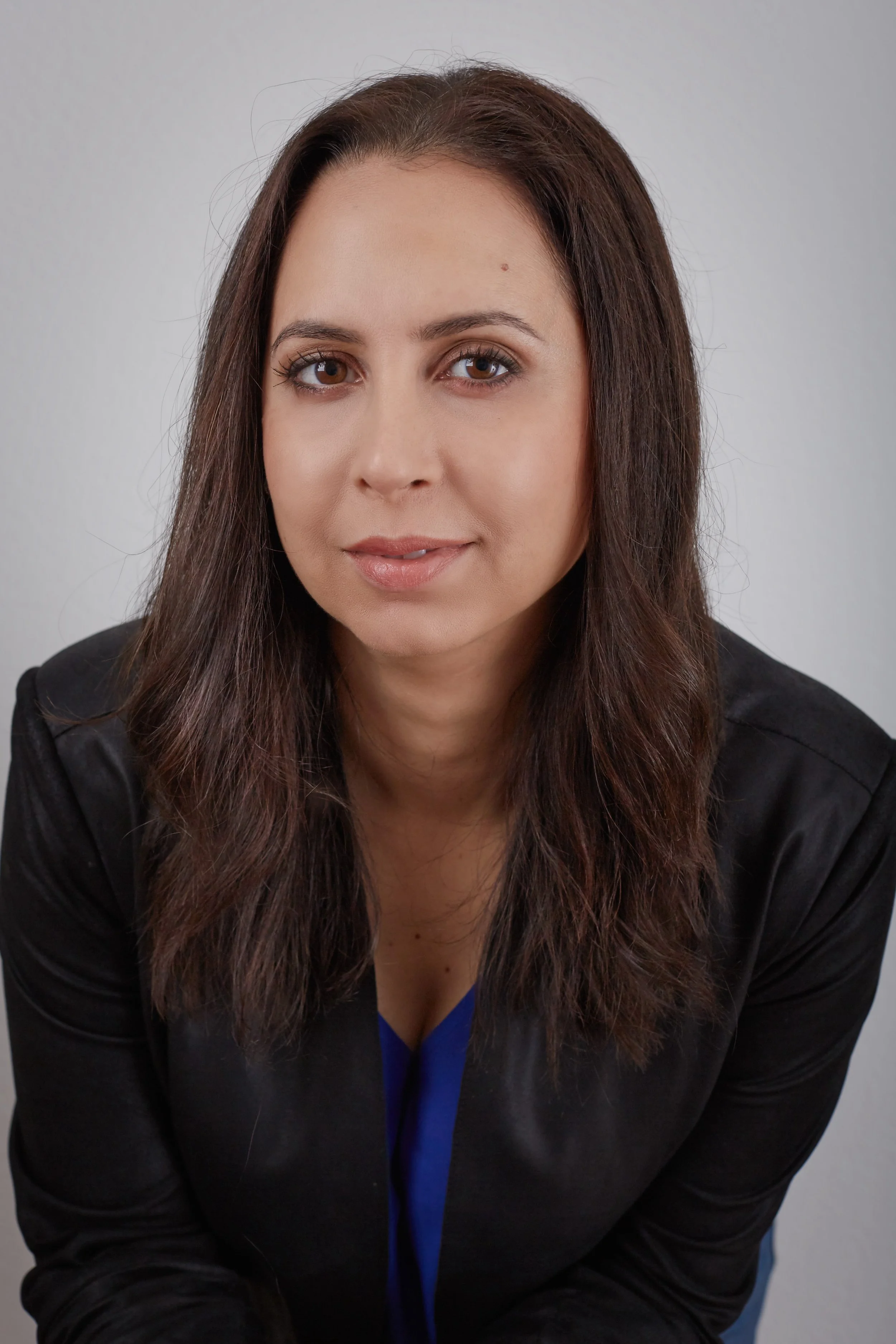 A woman with long brown hair, wearing a black jacket over a blue top, sitting against a plain gray background.