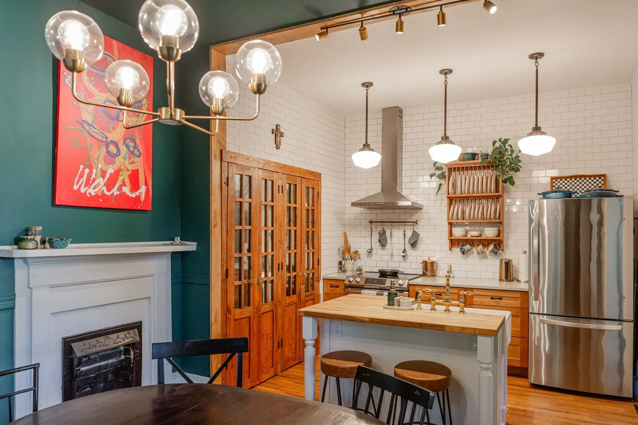 A cozy kitchen with white brick walls, wooden cabinets, and a refrigerator. There are three pendant lights hanging over the kitchen counter, a wooden dish rack, and a stove with cooking utensils. A small island with a wooden top has two stools. A colorful abstract painting hangs on a dark green wall near a fireplace.