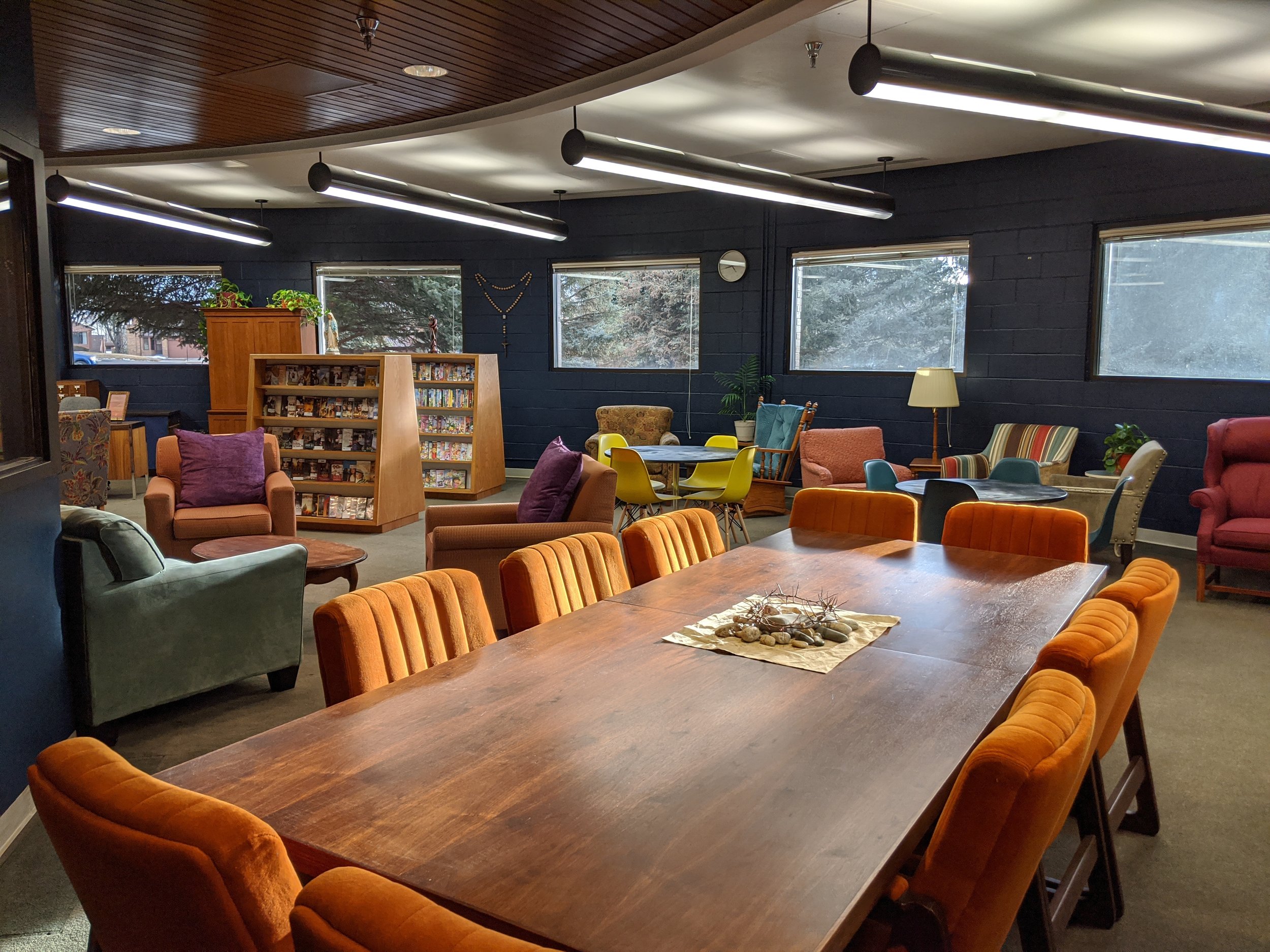 A room with dark blue walls, multiple windows, and a wood-paneled ceiling, furnished with colorful armchairs, a large wooden table, a bookshelf, plants, a lamp, and various decor items.