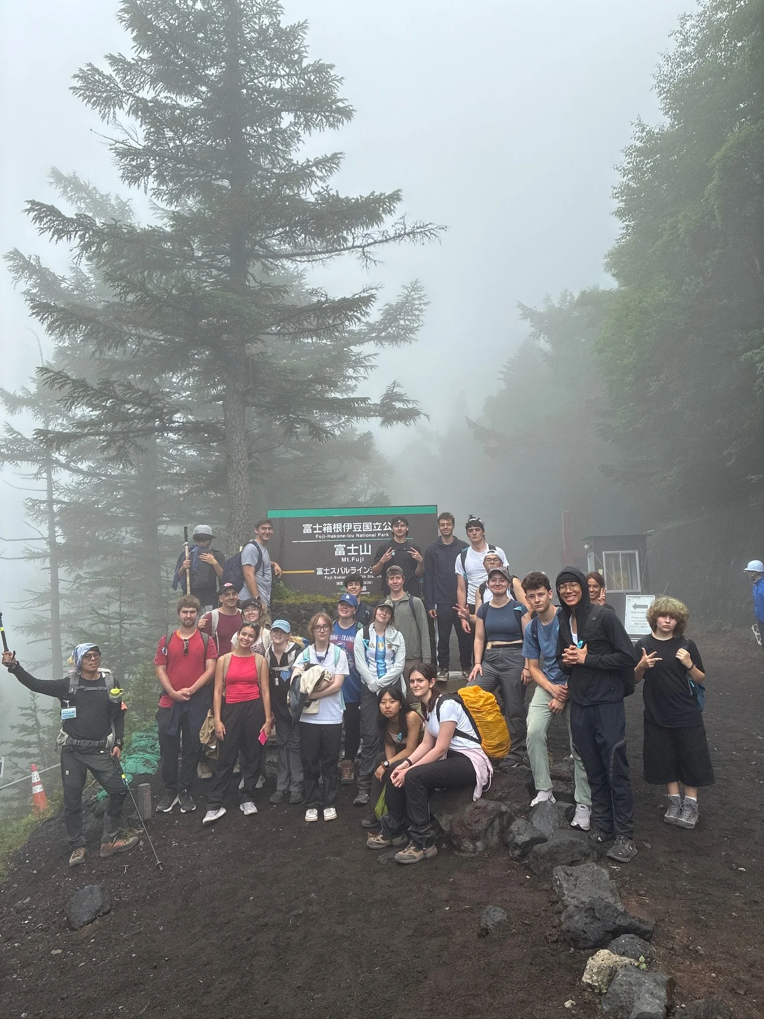 A large group of students hiking in a foggy forest, standing on Mt. Fuji and behind them tall evergreen trees.