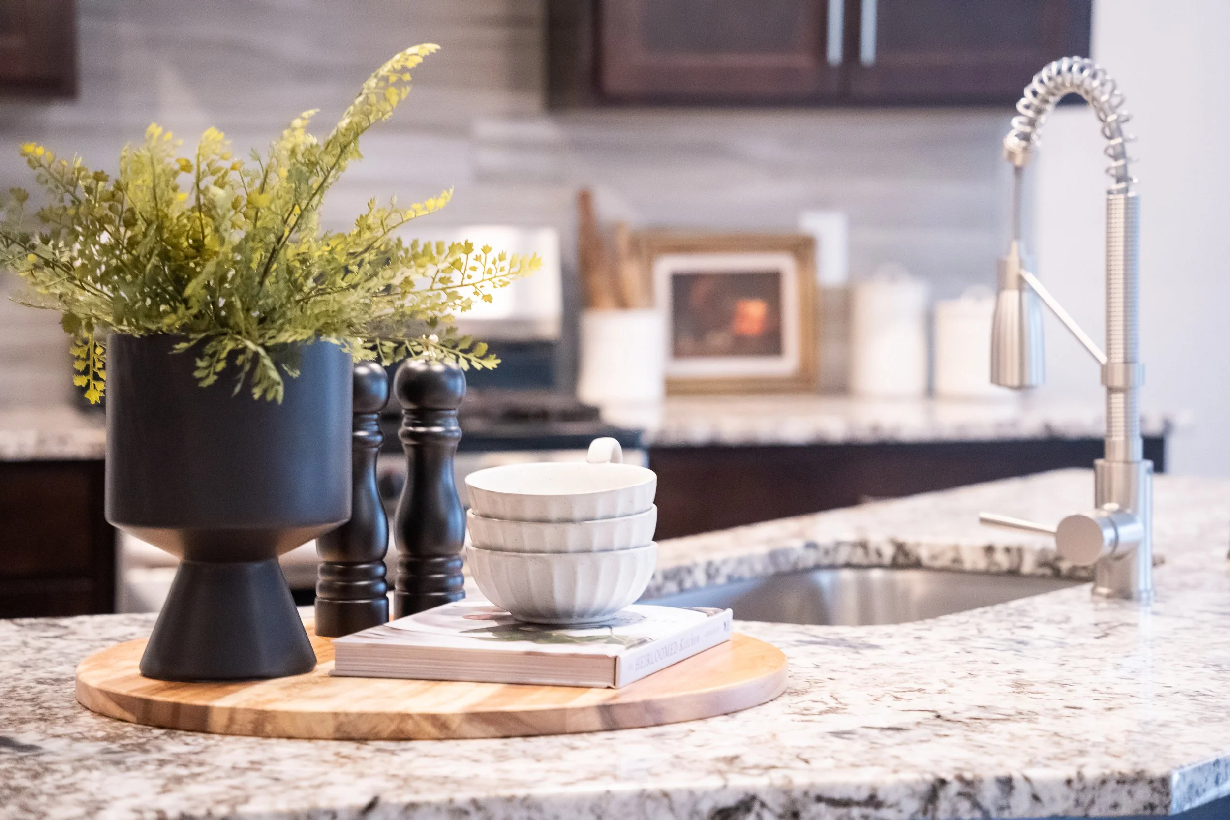 Staged kitchen countertop with wooden tray holding a black vase containing green artificial foliage, a pair of black salt and pepper shakers, and white bowls stacked on top of a book, next to a kitchen sink with a silver faucet.