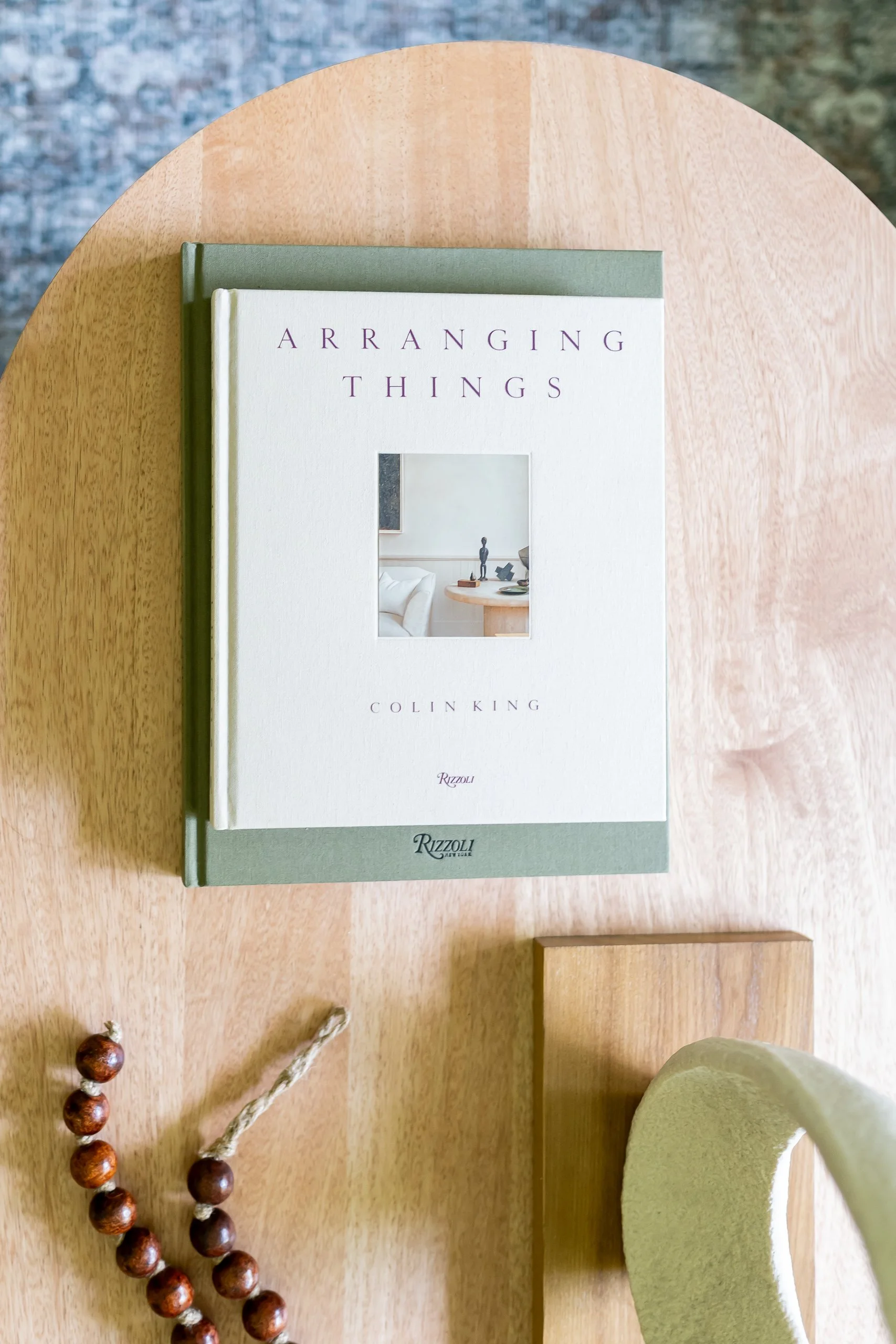 A hardcover book titled "Arranging Things" by Colin King on a round wooden table. Next to the table, there is a beige wall and a potted plant with a green, fuzzy leaf. A string of wooden beads is partially visible at the bottom left corner of the image.