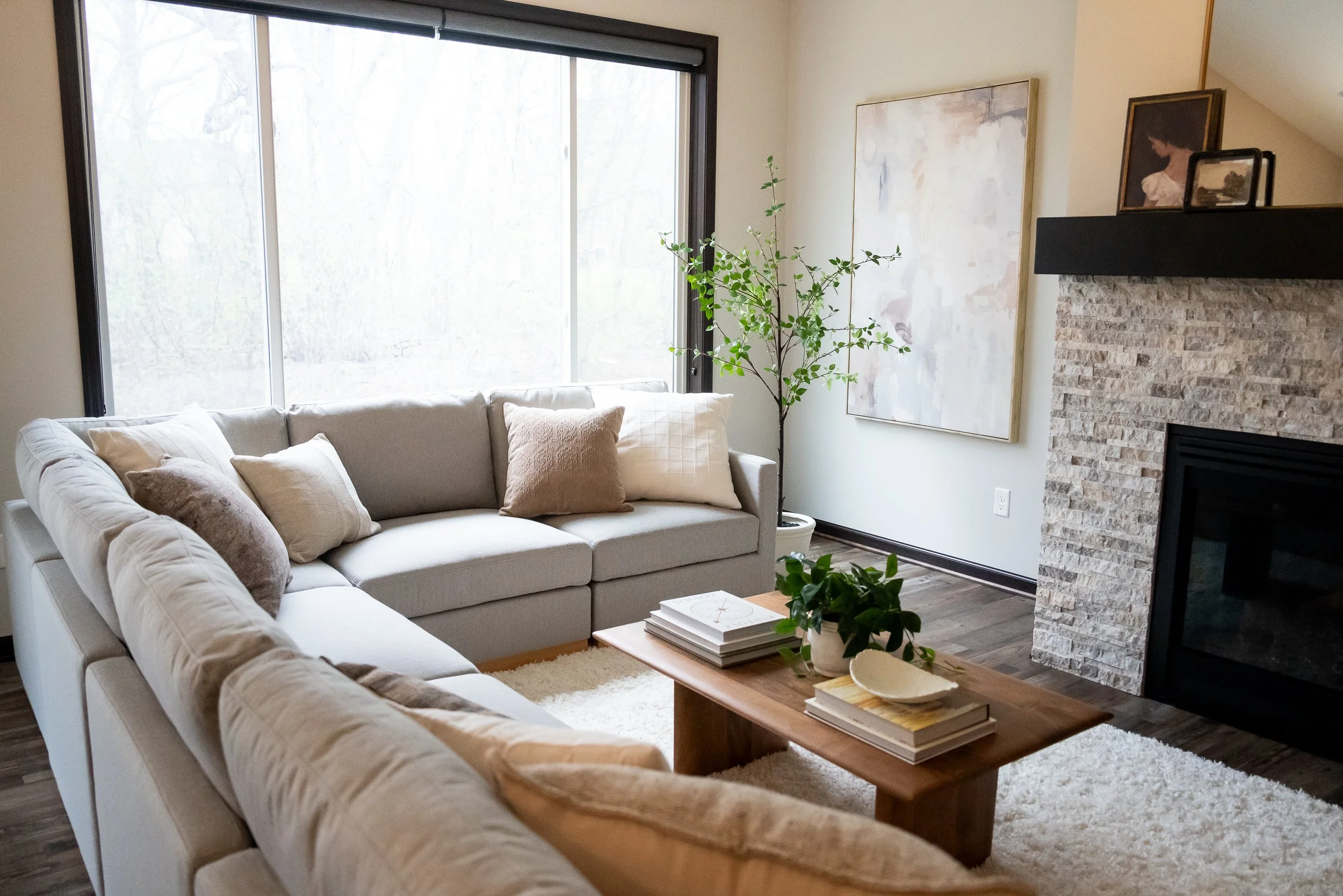 Staged living room with a large gray sectional sofa with various accent pillows, a wooden coffee table holding stacks of books and a plant, a fireplace with a stone surround displaying a mirror and small art, wall art, a tree and hardwood floors.