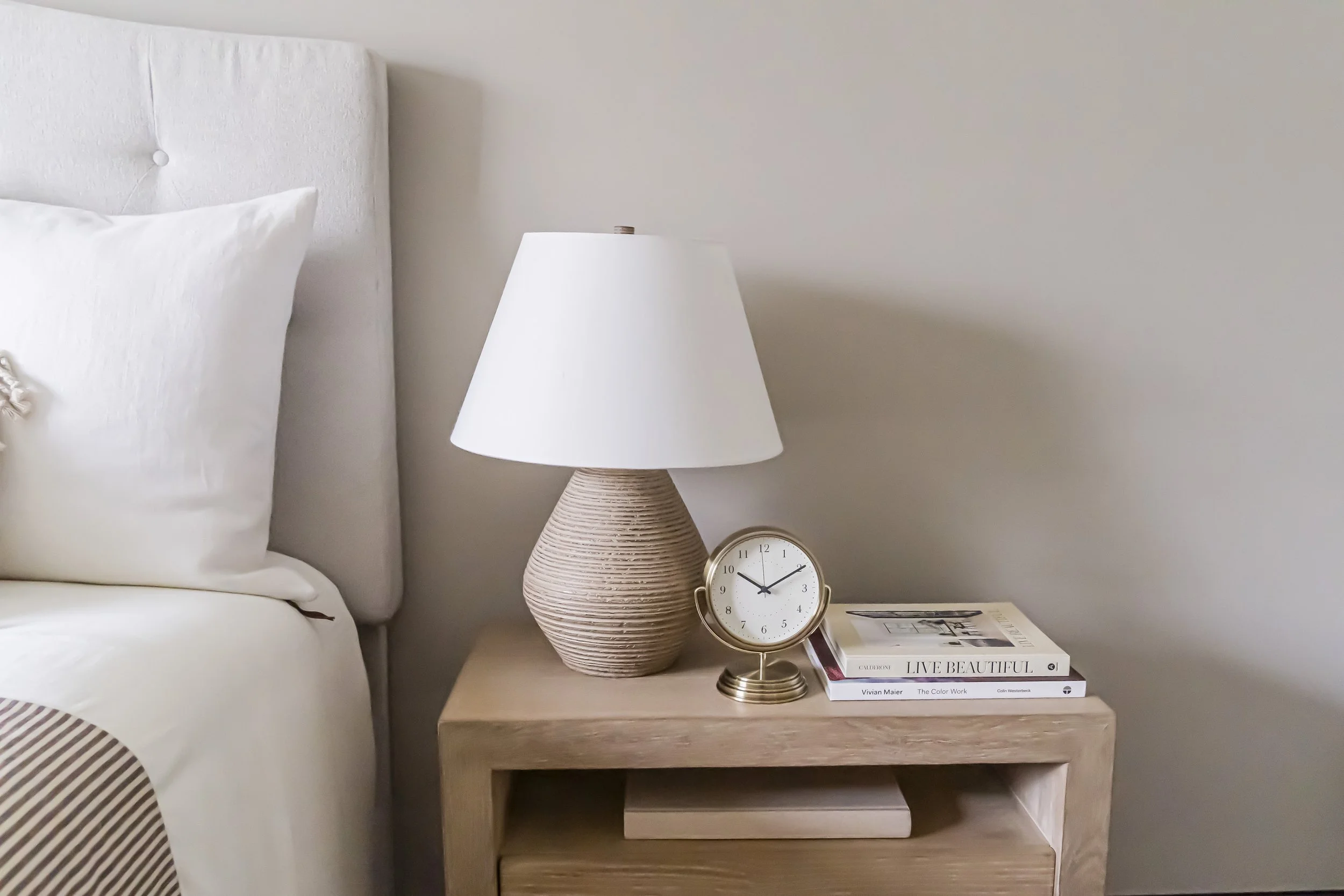 Staged bedroom nightstand with a beige textured lamp, a decorative clock, and a stack of books next to a bed with white linens and pillows, and a fabric headboard. The room has a light-colored wall.