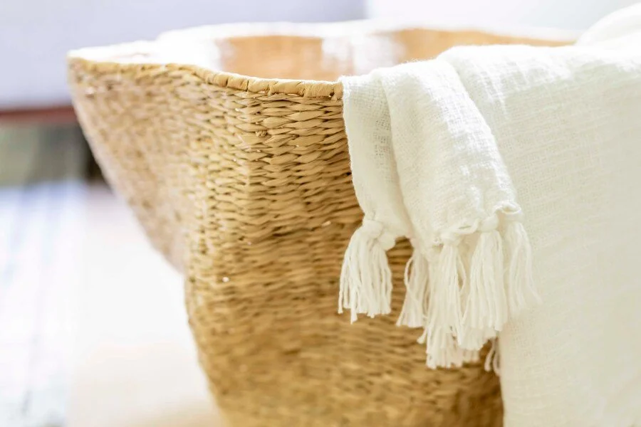 Close-up of a natural woven wicker basket staged with a cream tassel throw blanket draped over the side.
