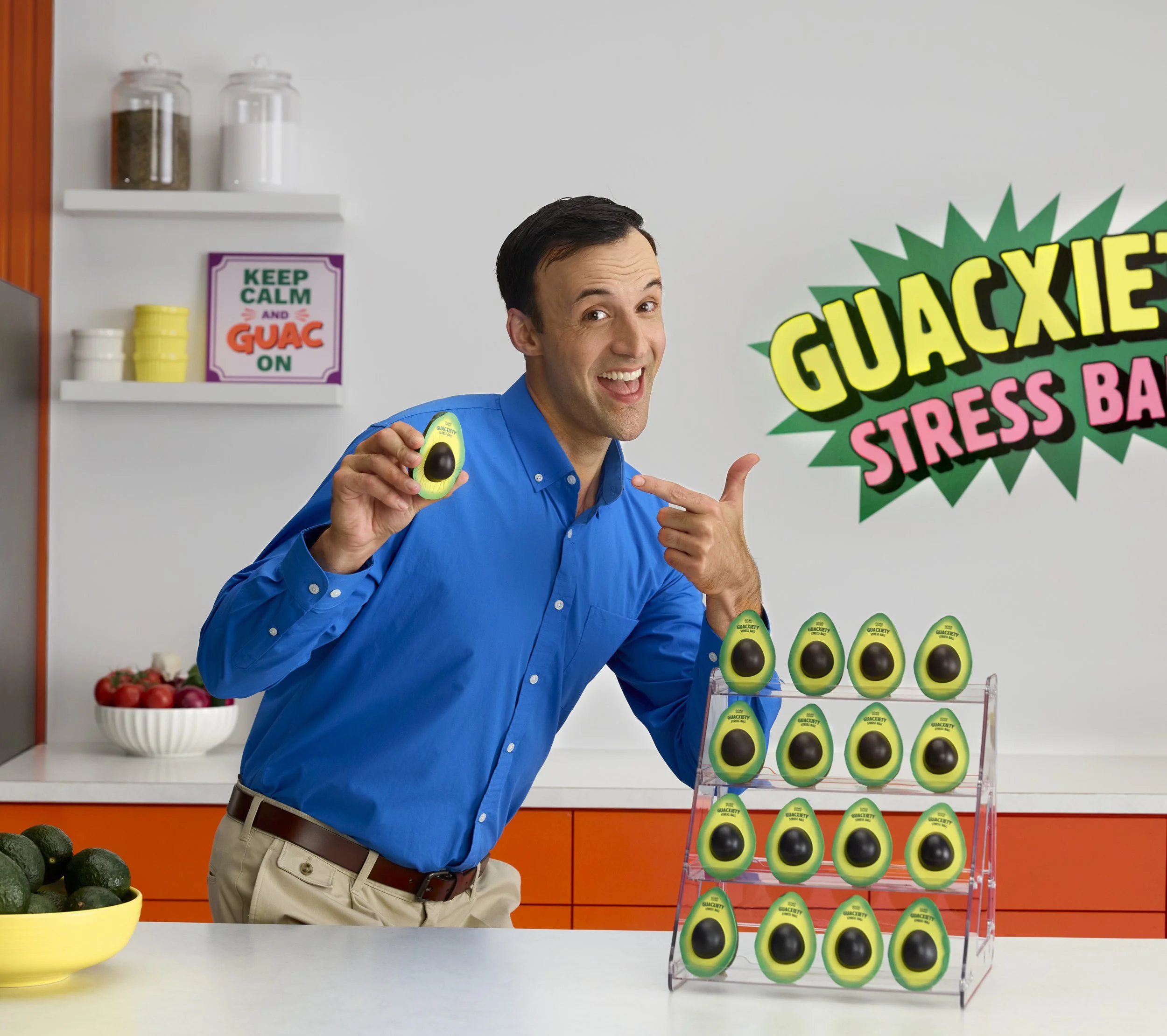 A man in a blue shirt points to a display of large avocado stress balls at a store counter. The background includes a sign that reads 'GUCAXE STRESS BALL' in colorful letters, a bowl of strawberries, and a shelf with jars of spices and bowls.