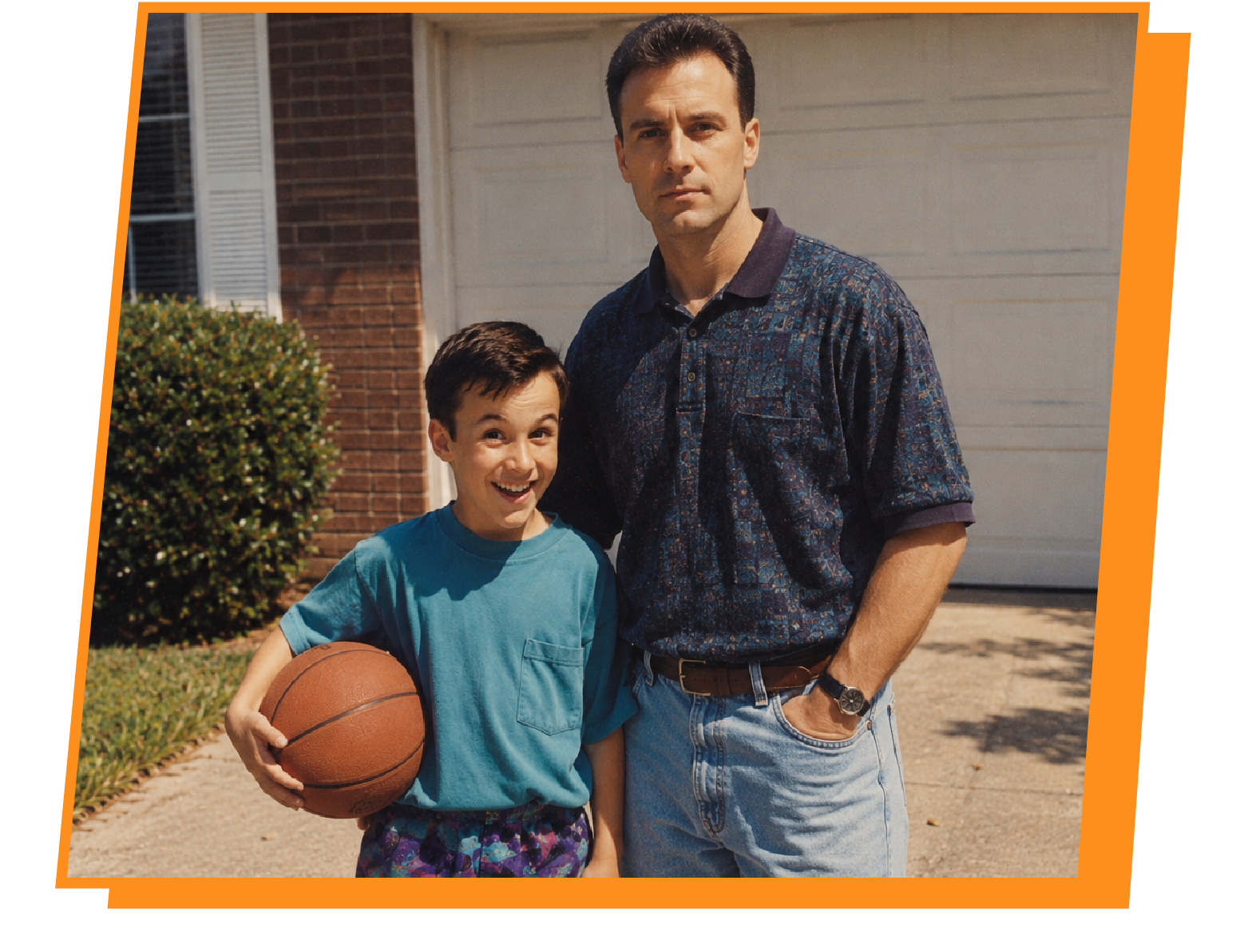 A young boy holding a basketball smiling next to an adult man, standing on a driveway in front of a house.