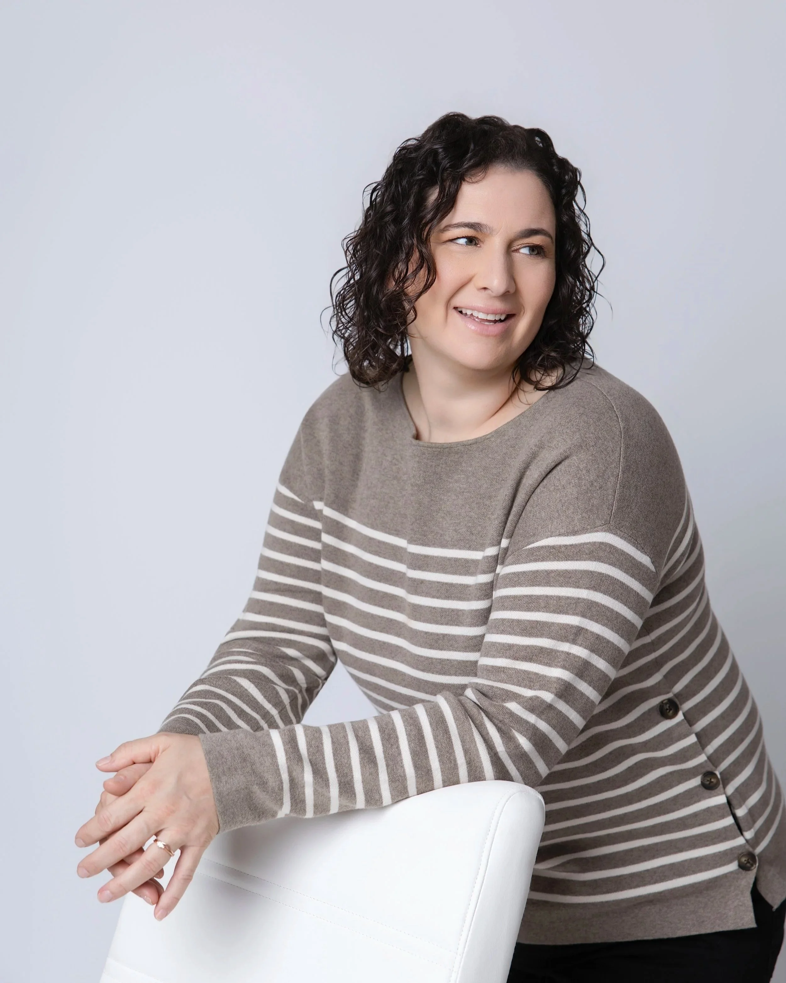 Jeanette smiling beside a white chair in a studio.