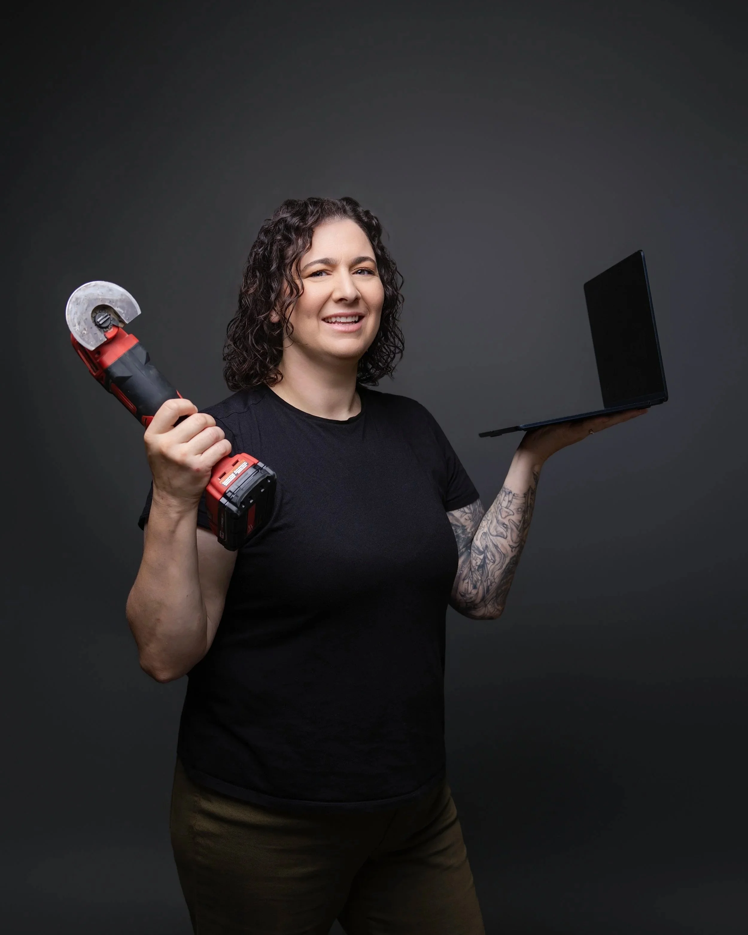 Jeanette holding a power tool and laptop against a dark studio background.