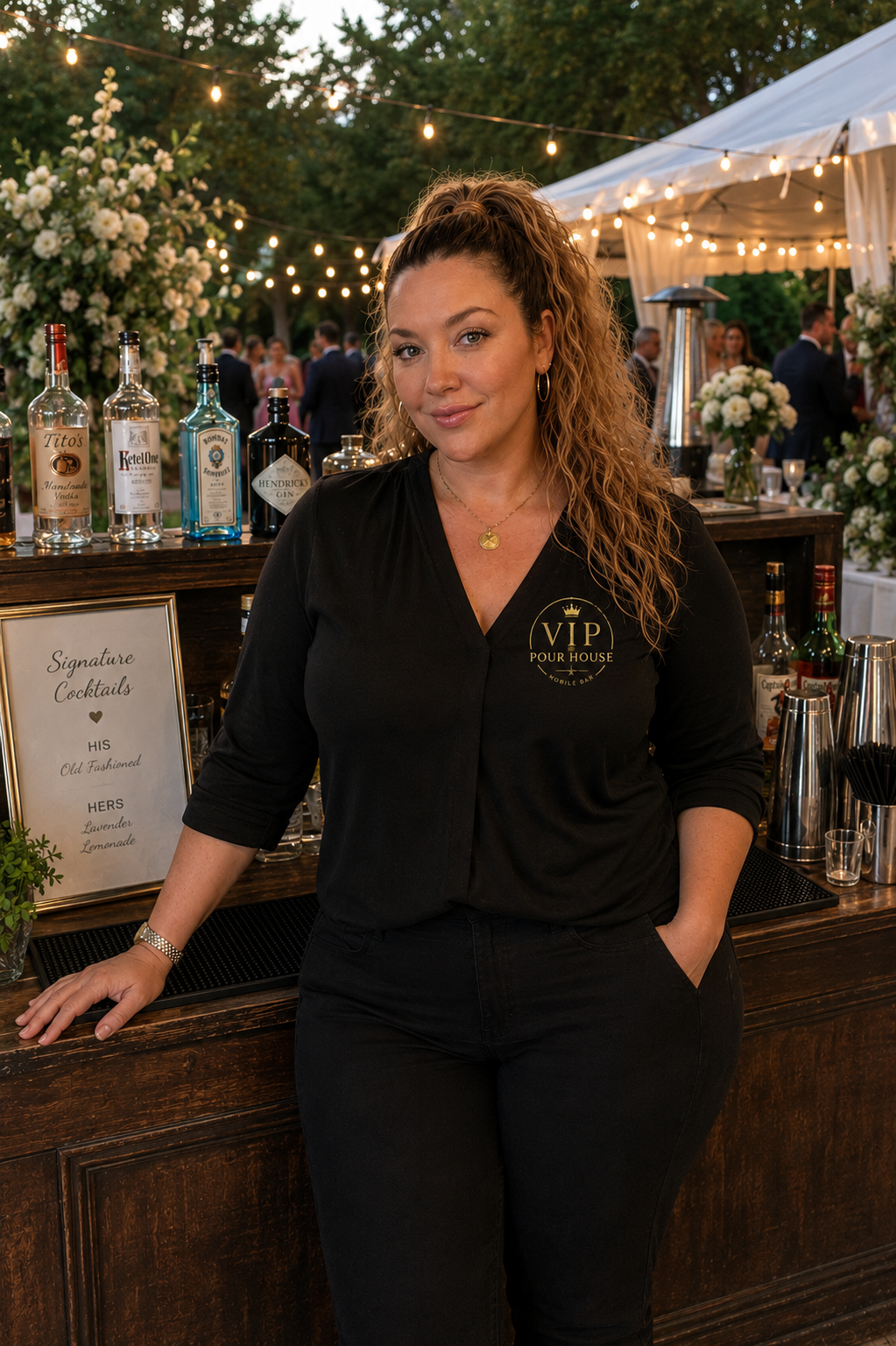 A woman with curly hair wearing a black shirt with 'VIP Pour House' logo poses at an outdoor event during the evening, with string lights and a bar setup behind her.