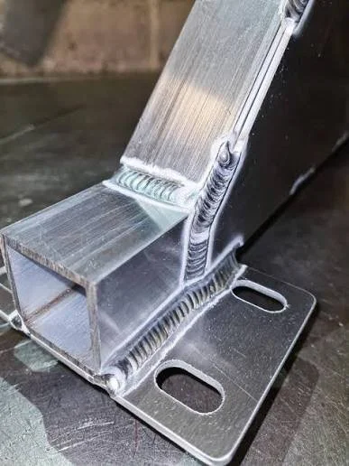 Metal bracket with welded joints, mounted on a flat metal base with two elongated holes, on a work surface.