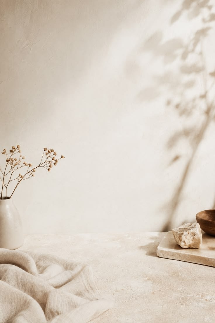 Minimalist interior with a beige textured cloth, a white vase with dried flowers, a marble slab with a stone, and a wooden bowl on a neutral-toned surface against a plain beige wall with soft shadowy foliage.