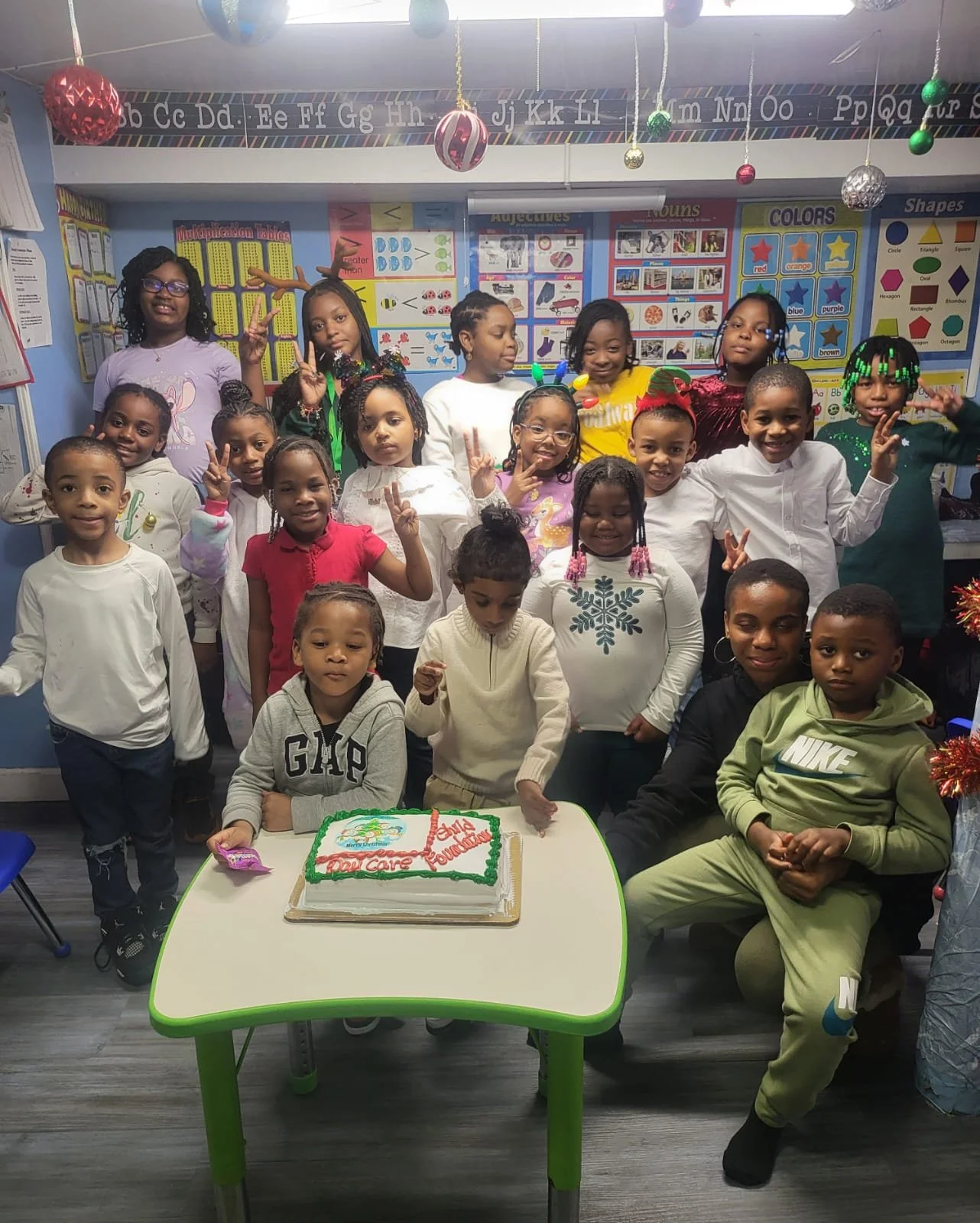A group of children in a classroom celebrating a holiday or special event, with a decorated cake on a table in front of them, and festive decorations hanging from the ceiling.