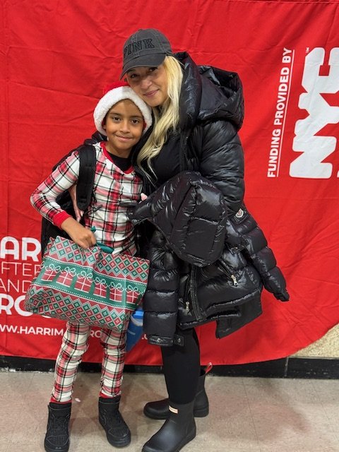 A woman and a girl standing in front of a red background, both wearing winter clothing and smiling. The girl is in Christmas pajamas, wearing a Santa hat, and carrying a large patterned gift bag. The woman is dressed in a black winter coat, black boots, and a gray cap.