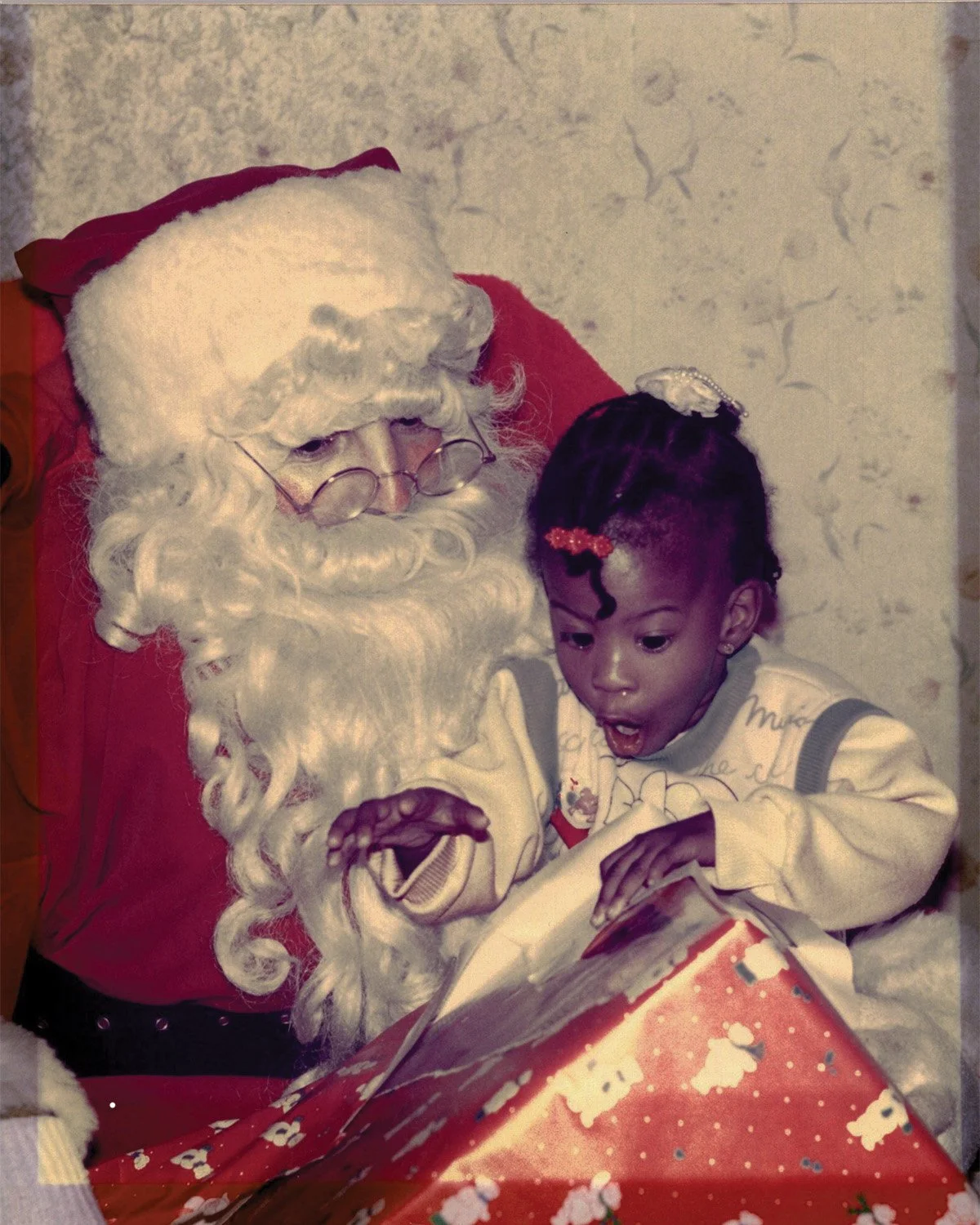 Santa Claus with white curly beard and glasses sitting next to a young girl with braided hair, holding a large wrapped Christmas present.