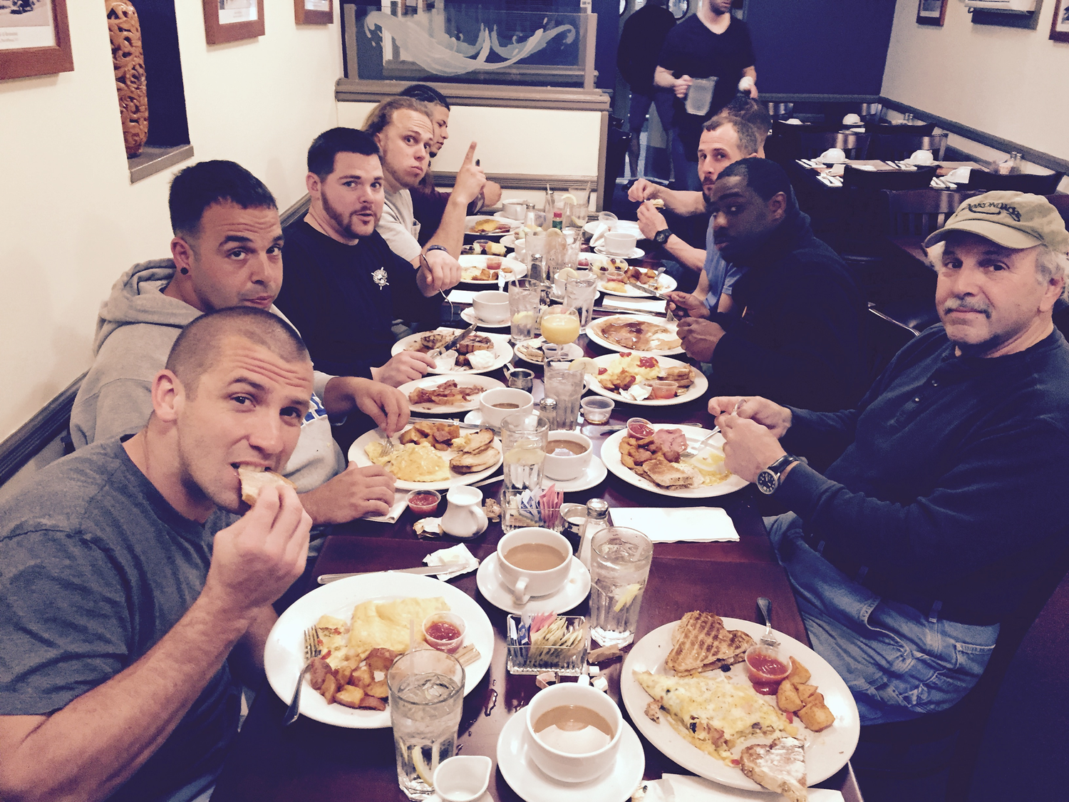 A group of nine men sitting around a long table in a restaurant, eating breakfast. The table has plates with waffles, pizza, scrambled eggs, and other breakfast items, as well as glasses of water and coffee.