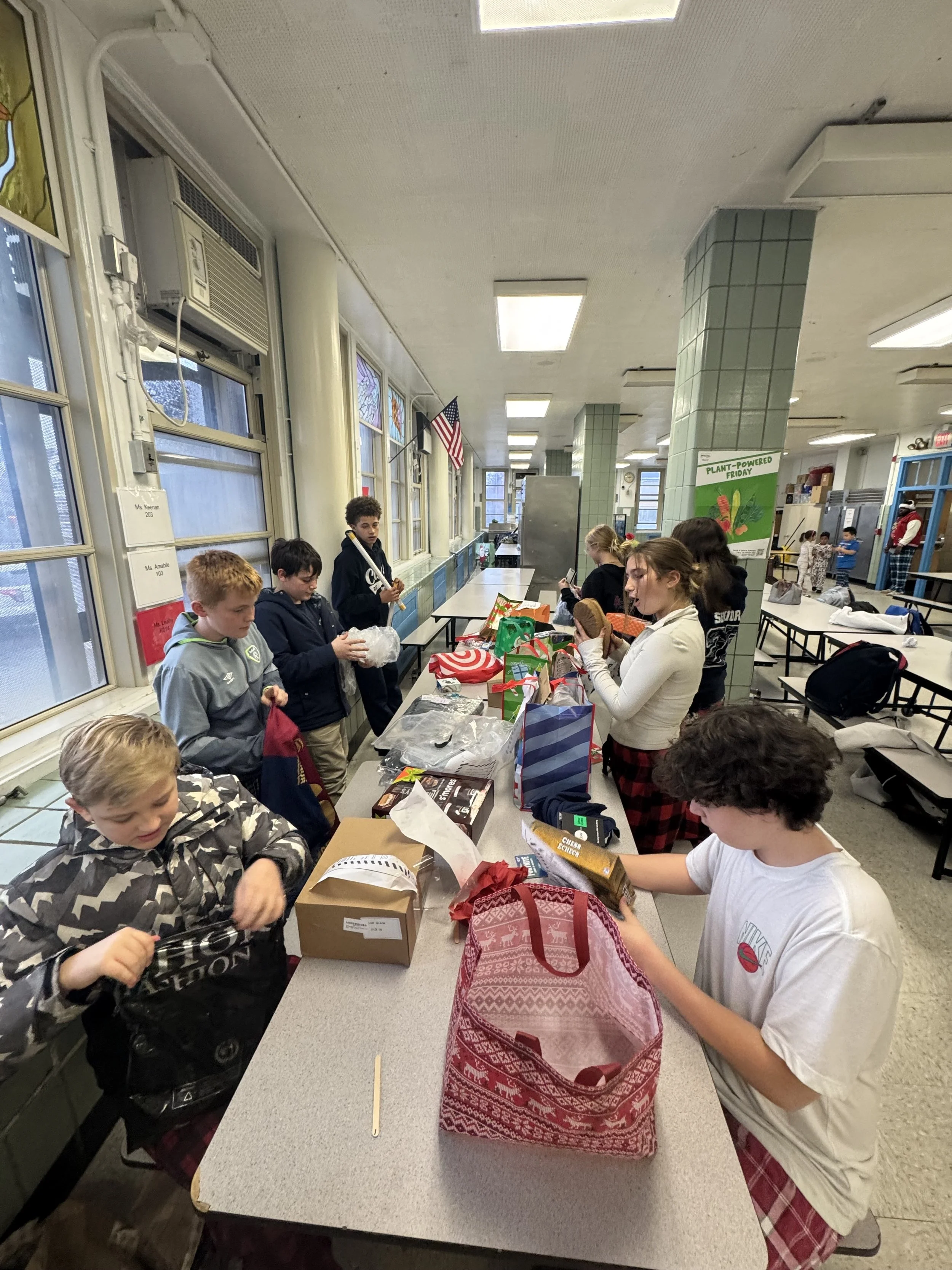 Children wrapped in winter clothing opening presents at a table in a school cafeteria decorated for Christmas.