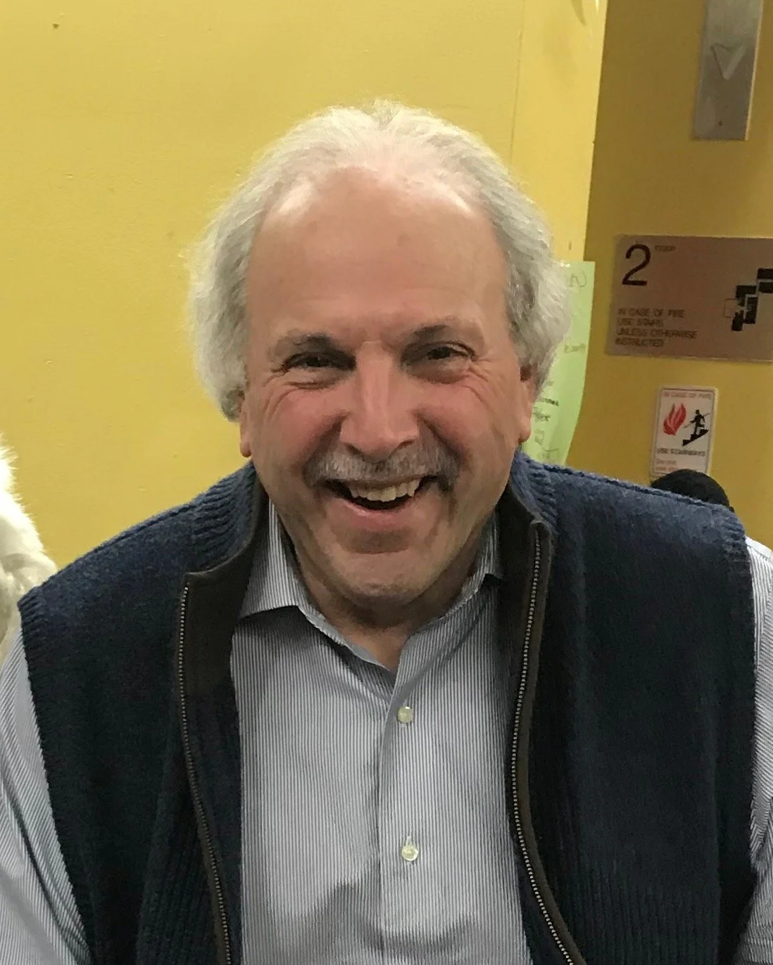 A smiling man with white hair and a mustache, wearing a striped shirt and a blue vest, standing in front of a yellow wall with signs and a fire extinguisher.