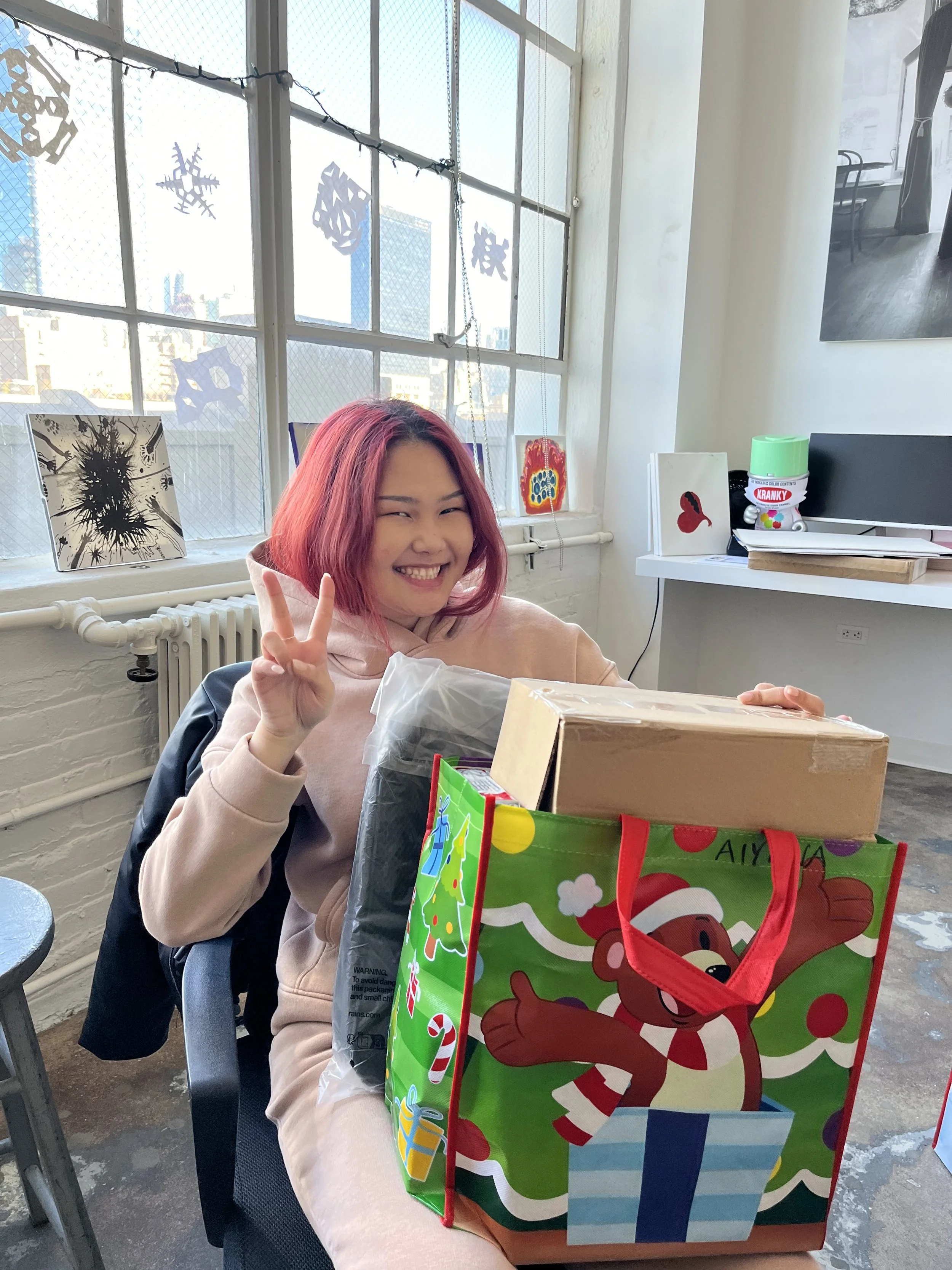 A young woman with pink hair smiling and making a peace sign while sitting at a table with Christmas presents, a colorful gift bag with a reindeer, and a window decorated with snowflake cutouts.