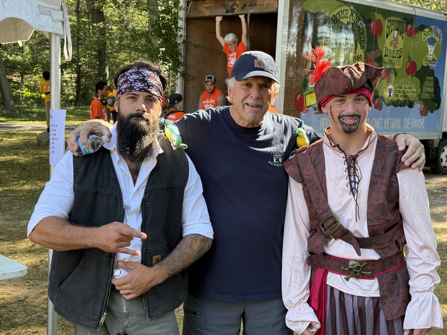 Three men dressed as pirates posing together at an outdoor event with a food truck and other people in the background.