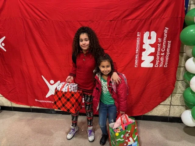 Two young girls stand in front of a red backdrop with white lettering and balloons on the right side. The older girl has long curly hair, wearing a red sweater, patterned leggings, and sneakers, holding a gift. The younger girl has long dark hair, wearing a green shirt, pink jacket, jeans, and boots, holding a colorful gift bag.