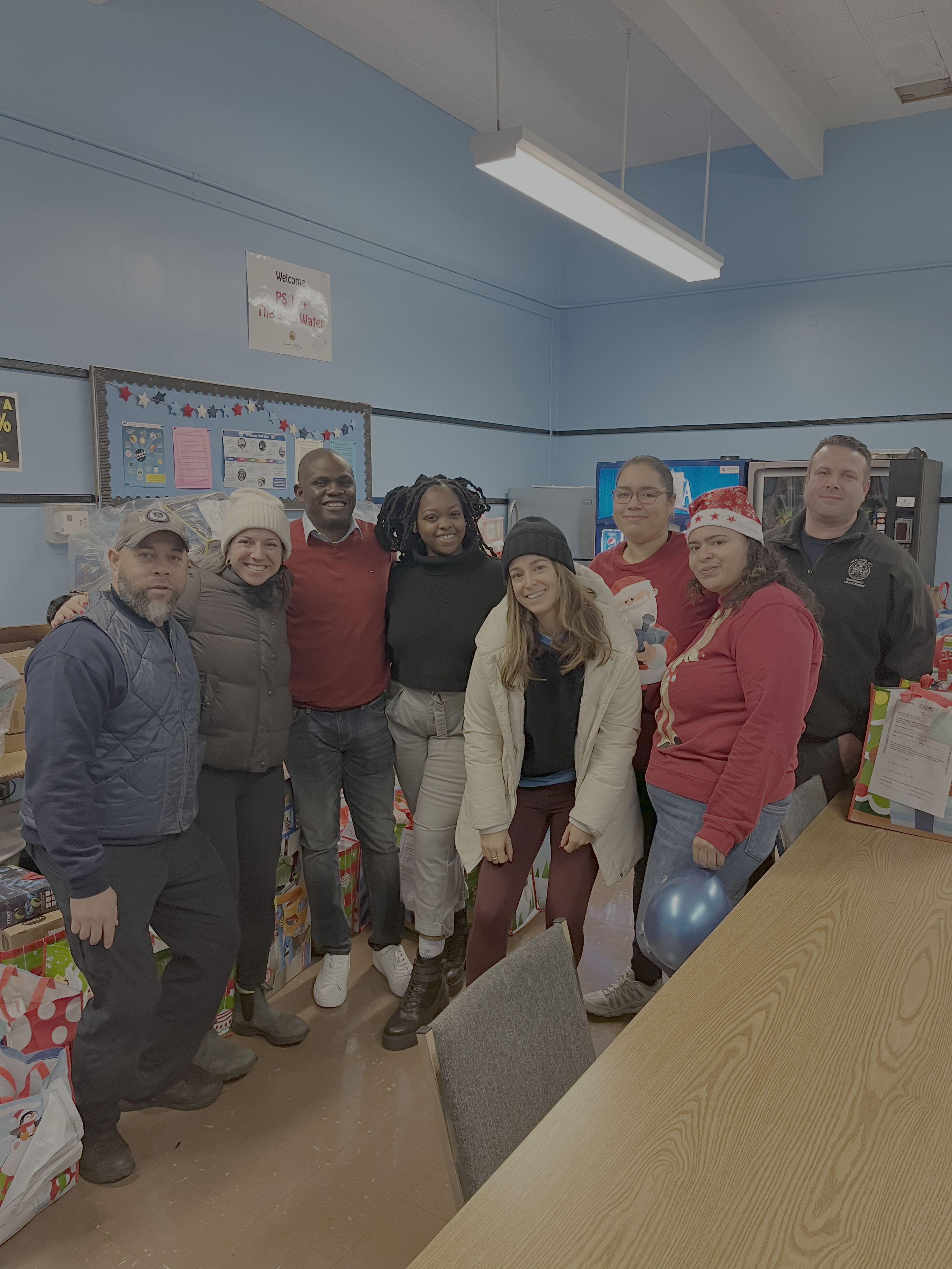 Group of people gathered together in a room, some wearing Christmas-themed apparel, smiling for a group photo, with gift bags and holiday decorations in the background.