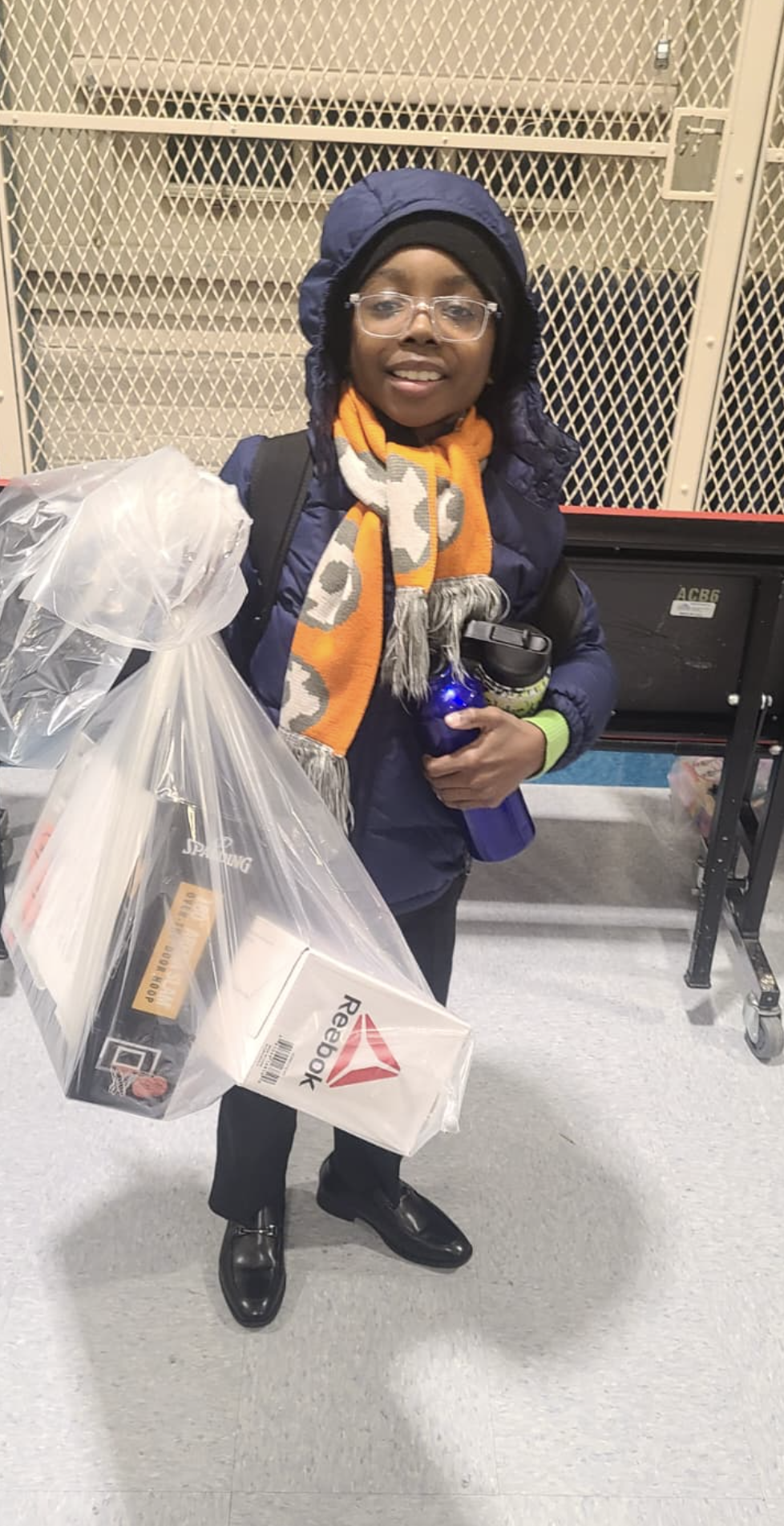 Person wearing glasses, a black hat, and a navy jacket, holding a large plastic bag filled with Reebok-branded items, standing in front of a wire mesh fence at an airport or similar transportation hub.