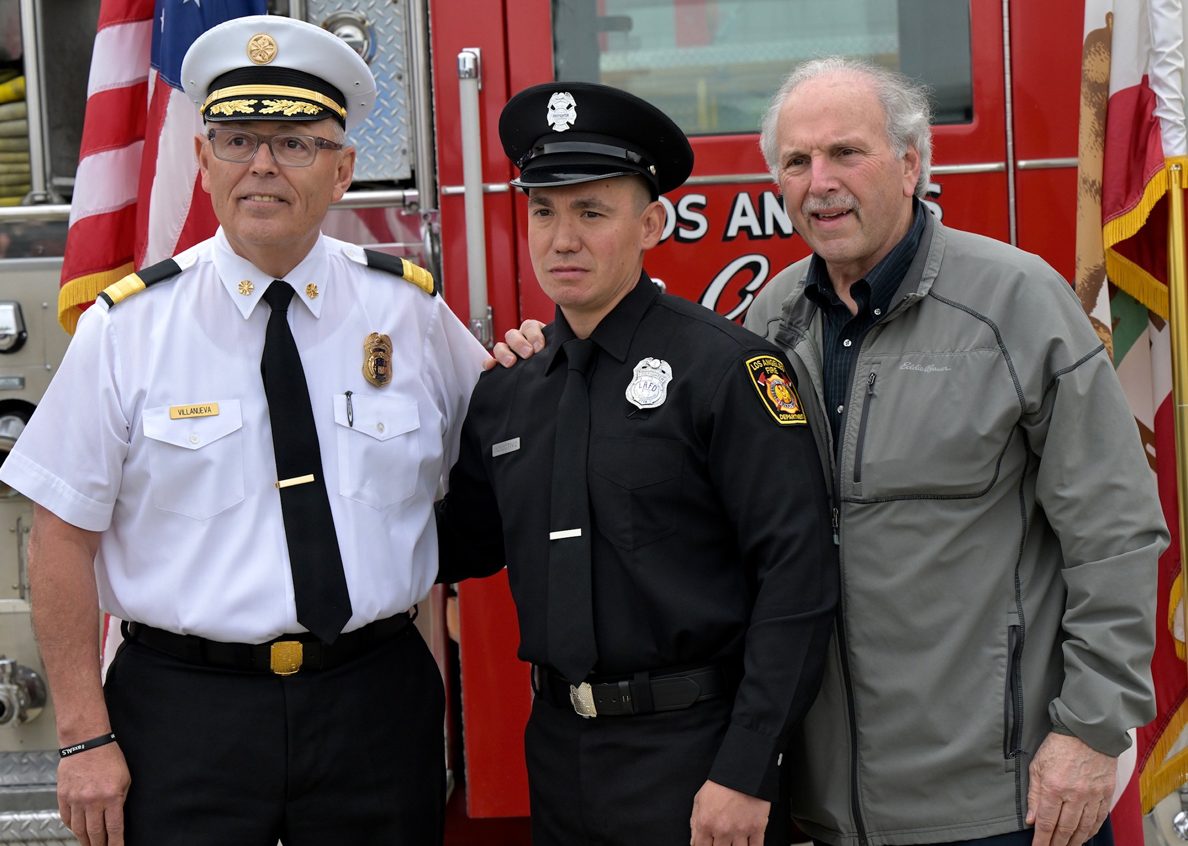 Three men standing in front of a fire truck, two of them in firefighter uniforms and the third in casual clothing, posing for a photograph.