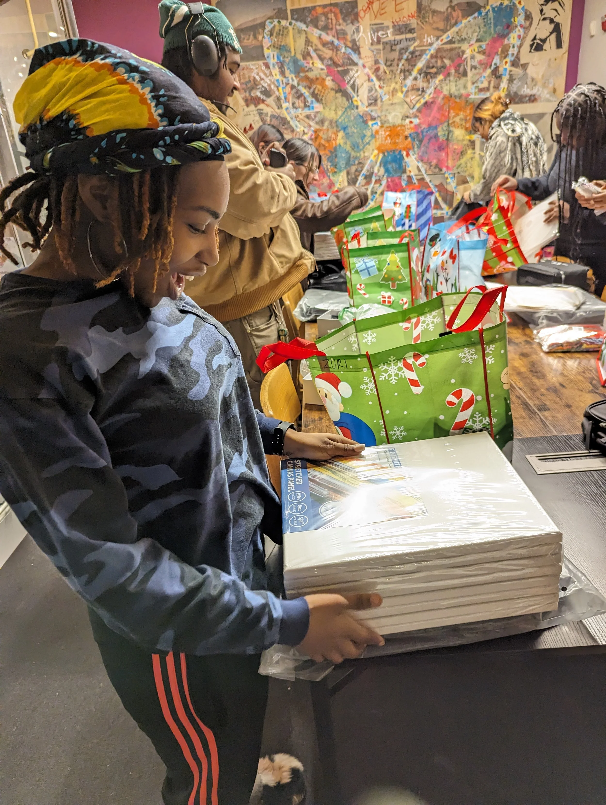 A group of people at a table wrapped in Christmas gift bags, opening presents, with a colorful mural on the wall behind them.