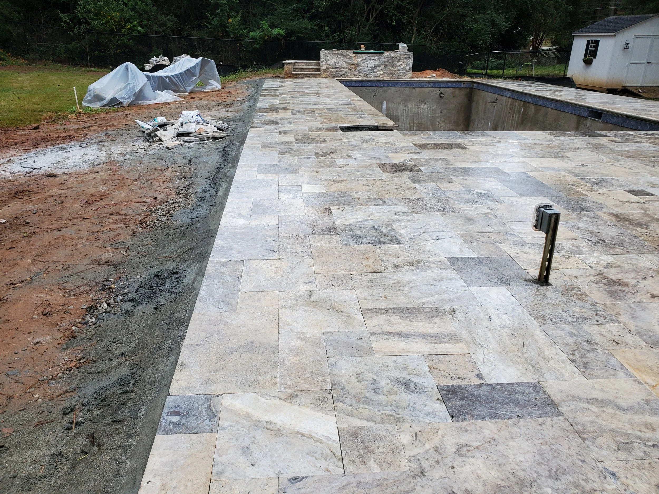 Construction site with newly laid stone tiles in a backyard, partially covered area with construction materials, and a swimming pool under construction.