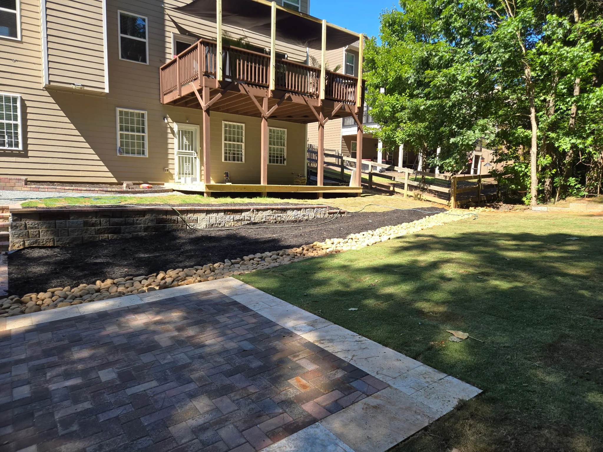 A backyard with a wooden deck extending from the back of a beige house, with a partially constructed railing. The yard has trees, grass, and a stone-bordered garden bed with mulch. There is a paved patio area with a brick pattern in the foreground.