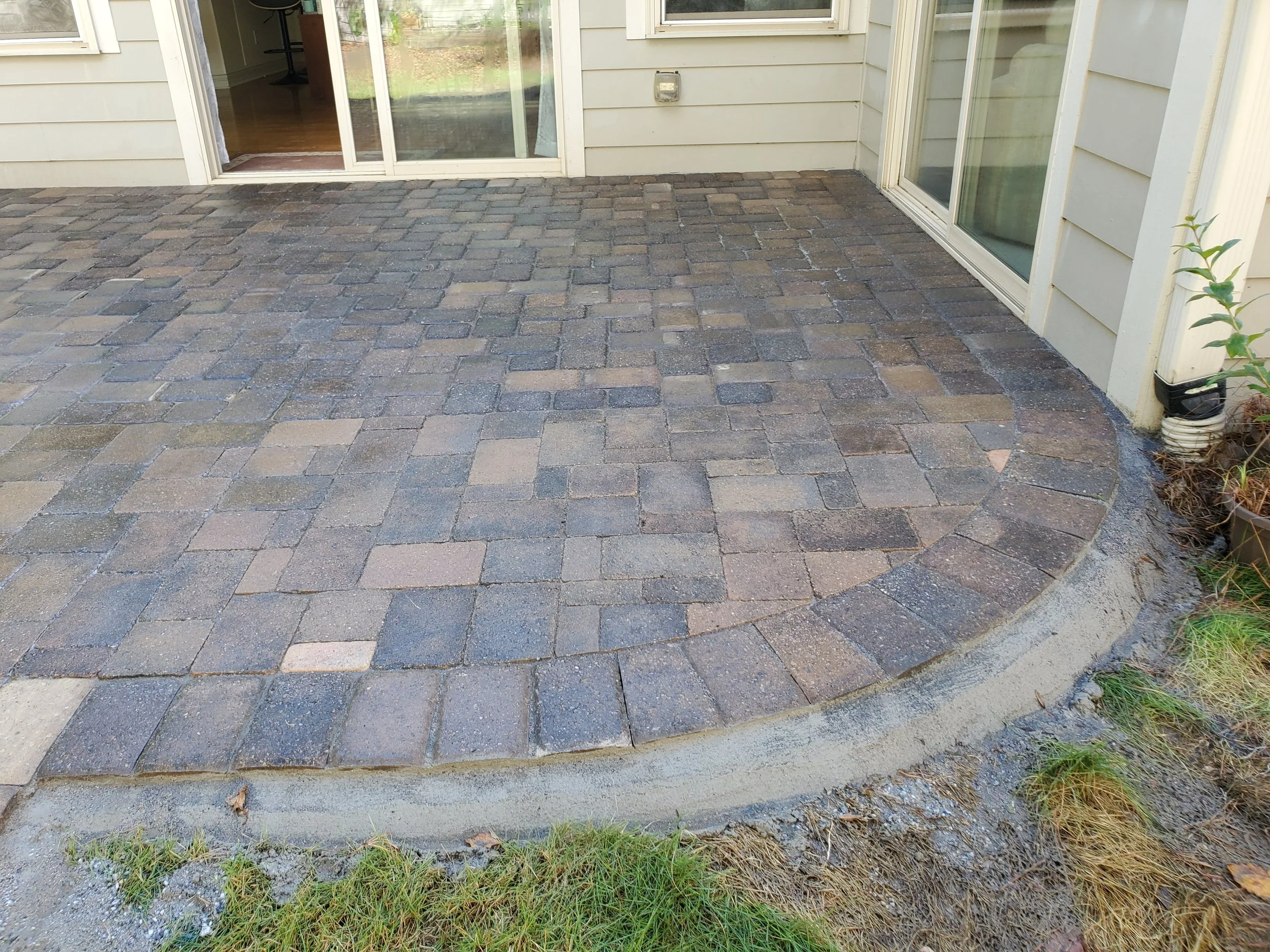 A patio with brick paving stones outside a house with sliding glass doors and beige siding. There is a small plant on the right side and a patch of grass at the edge.