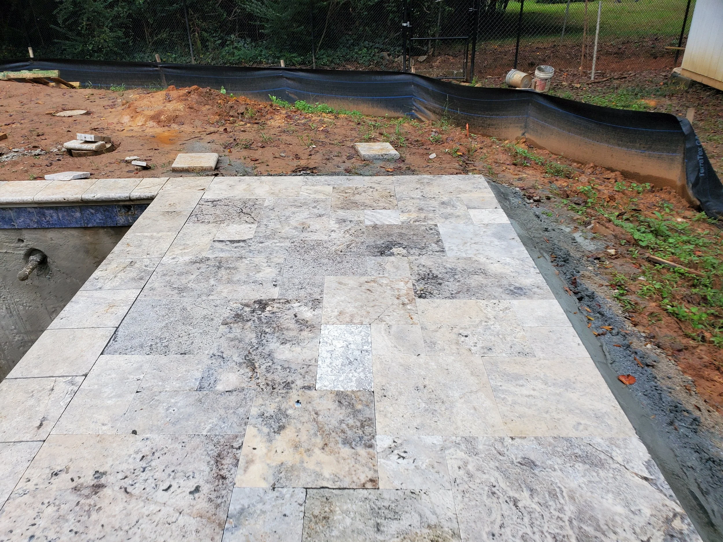 A backyard area with a partially completed stone patio and a garden bed with dirt, some small plants, and construction materials, including buckets and a black fabric barrier.