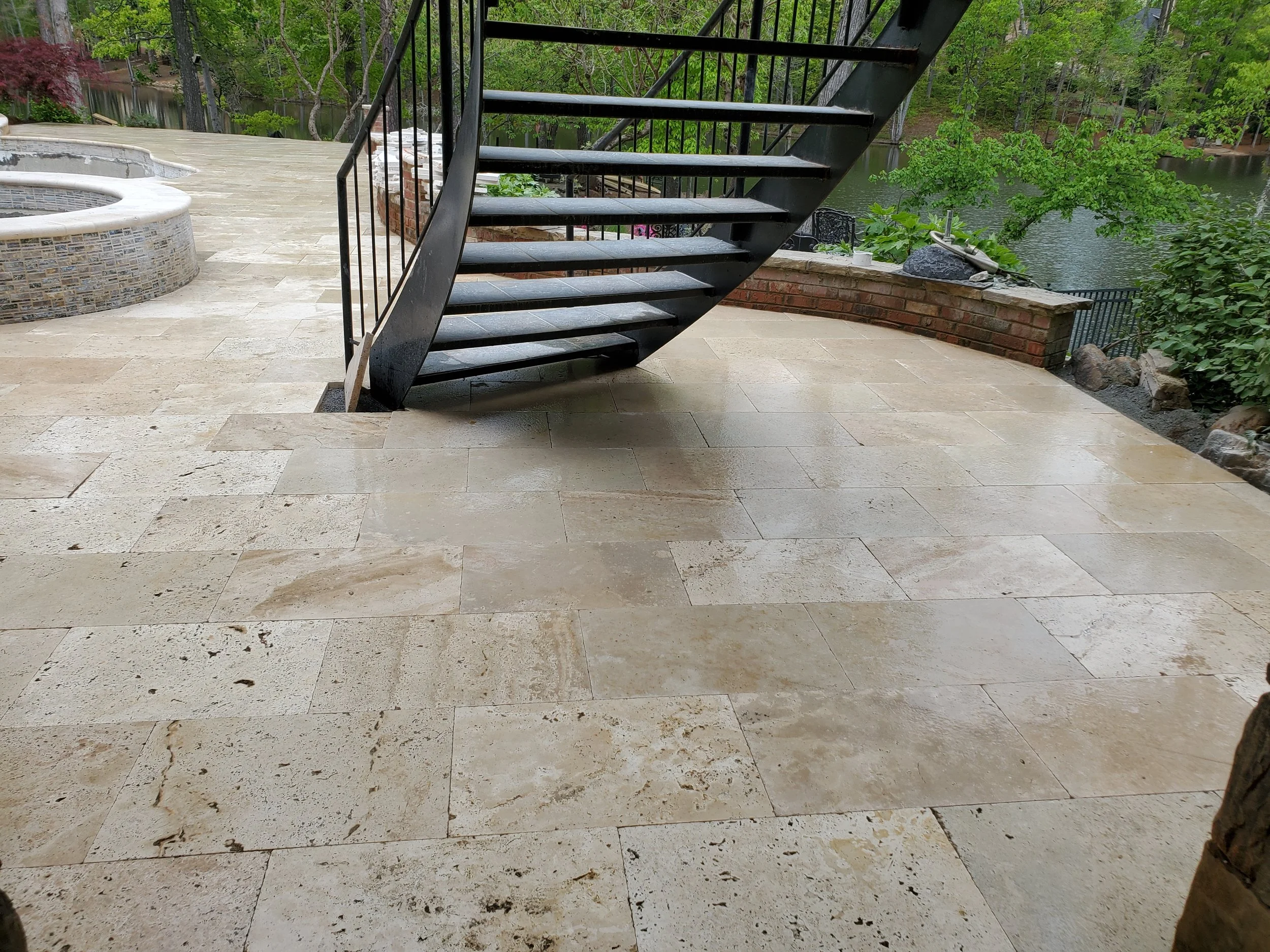 A backyard patio with beige stone tiles, a brick firepit, and a black metal staircase leading to a higher level, with greenery and a river in the background.