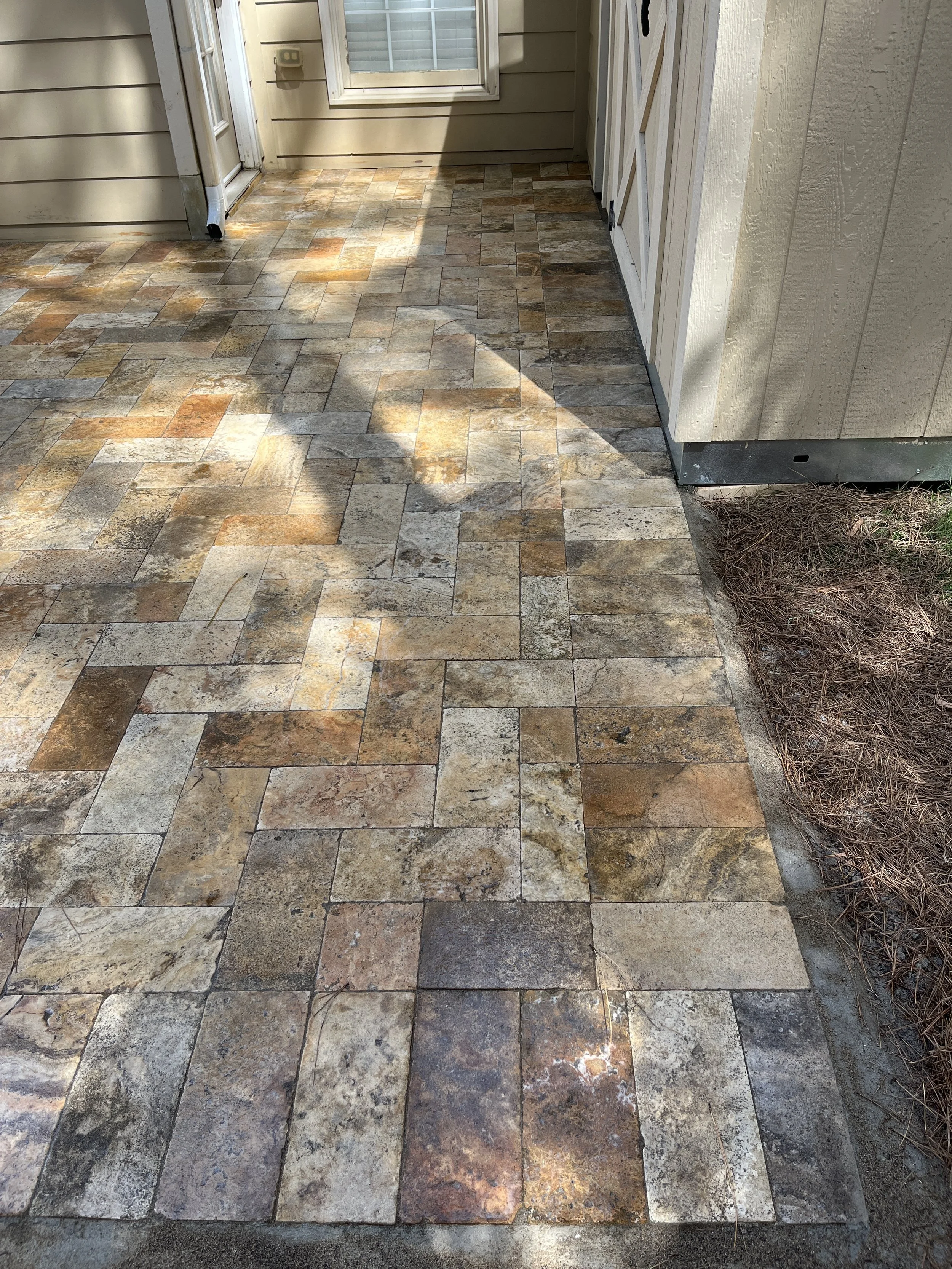 Close-up view of a backyard porch area with multicolored stone or tile paving, a beige siding house exterior, a window, a door, and some garden soil with dry pine needles on the right side.