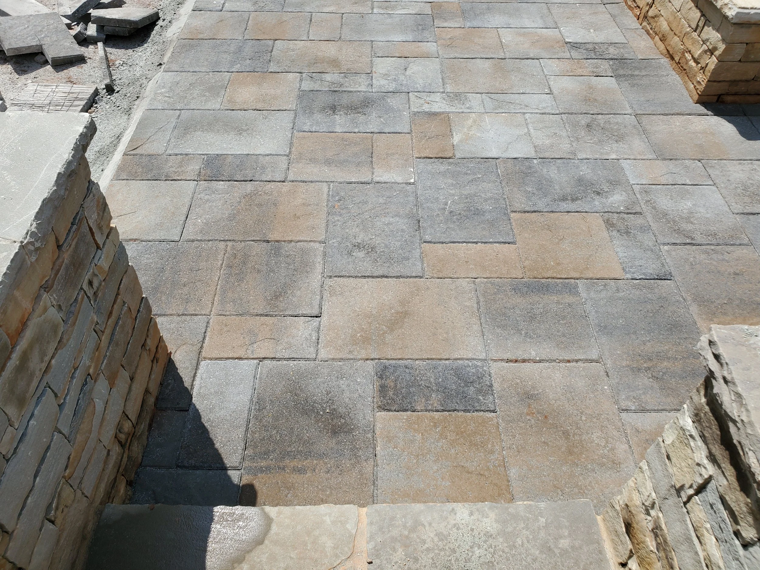 A newly paved outdoor patio area with rectangular stone pavers in shades of gray and tan, bordered by a brick wall on the left and right sides.