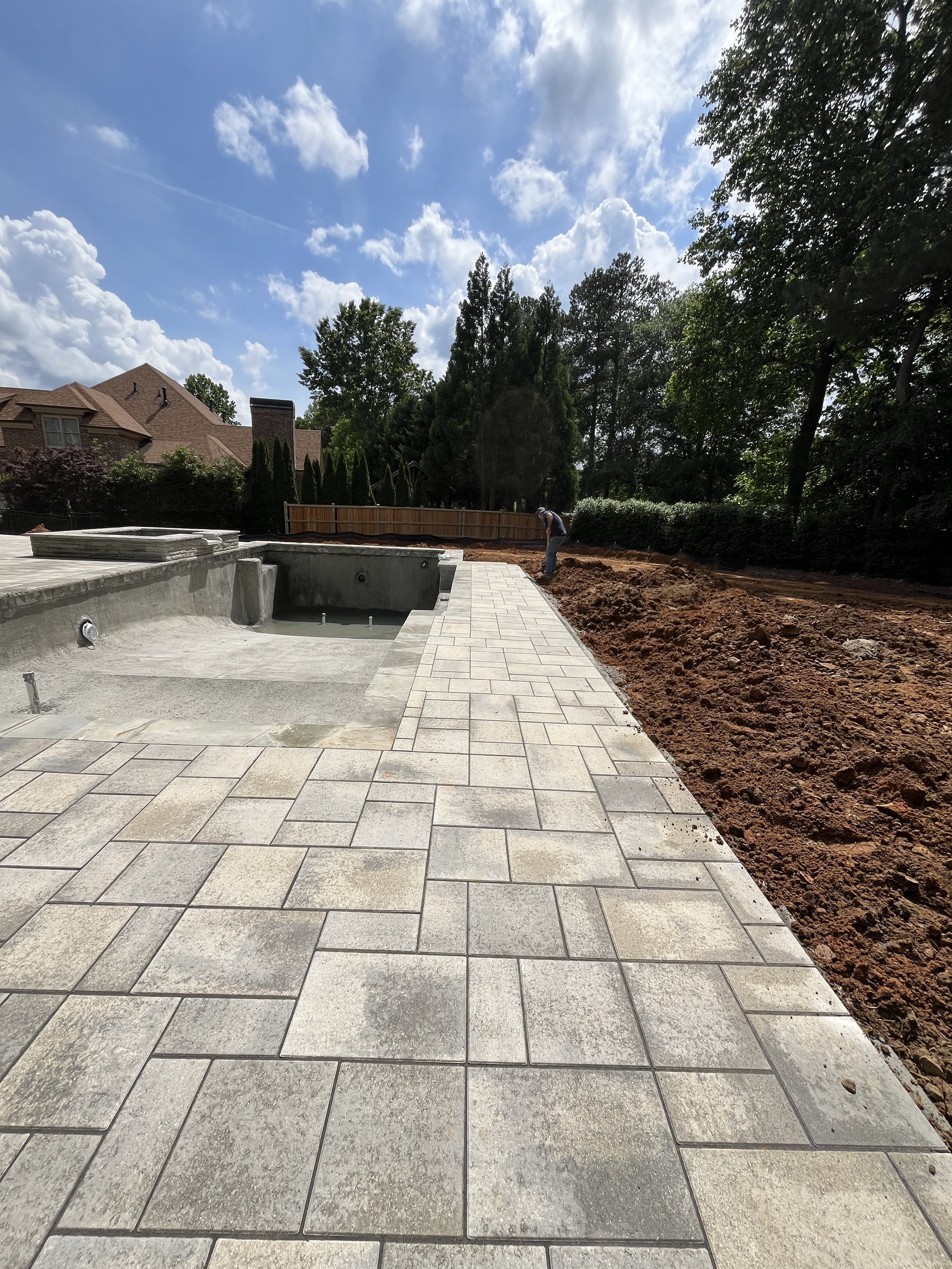 A backyard with a pool in the process of being built, with paving stones surrounding it, a worker laying down new pavers, and a wooden fence in the background under a partly cloudy sky.