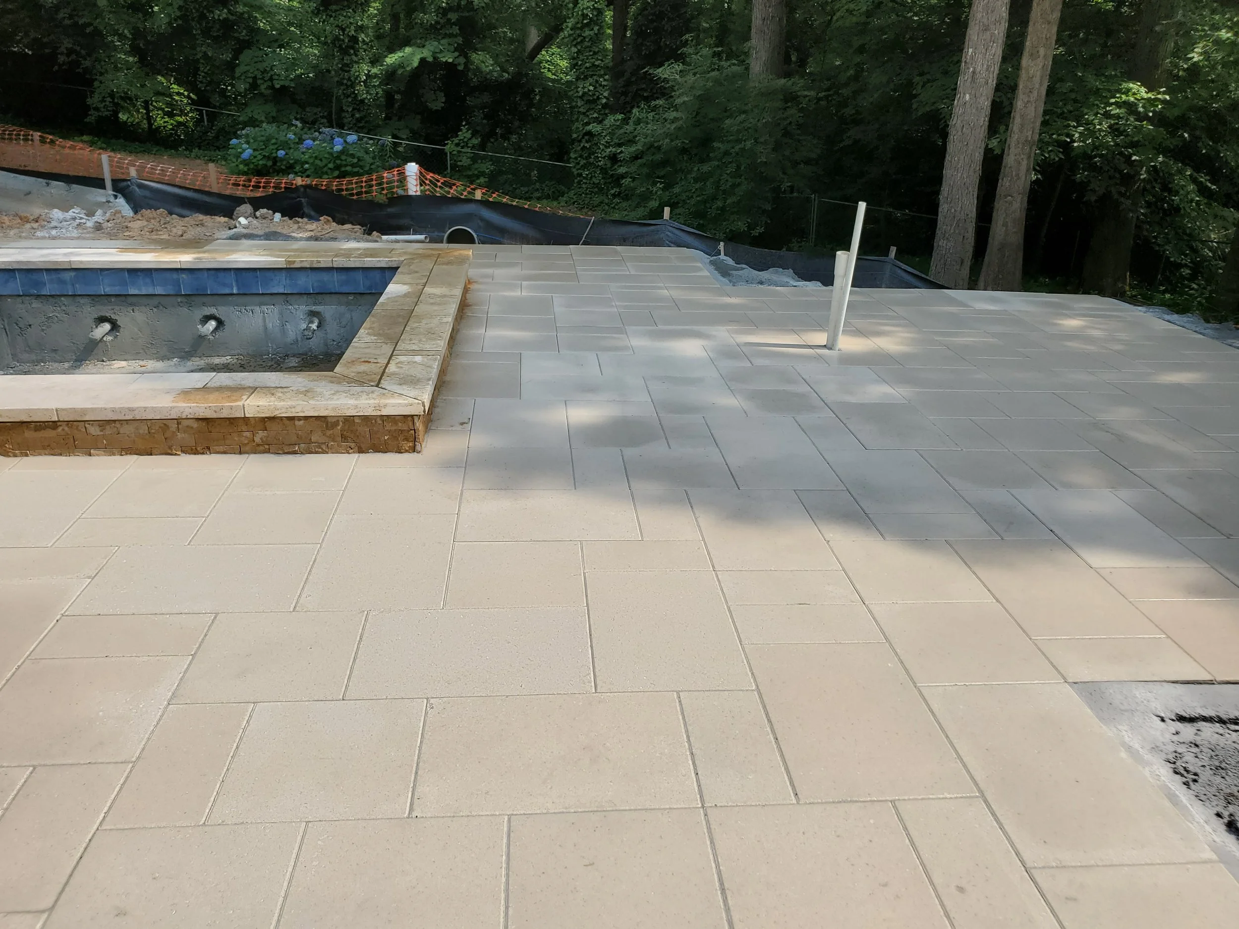 An outdoor scene showing a partially completed patio or deck with large beige tiles, a small built-in hot tub or pool with blue tiles, and a wooded background with green trees and shrubs. There are some construction materials and dirt visible, along 