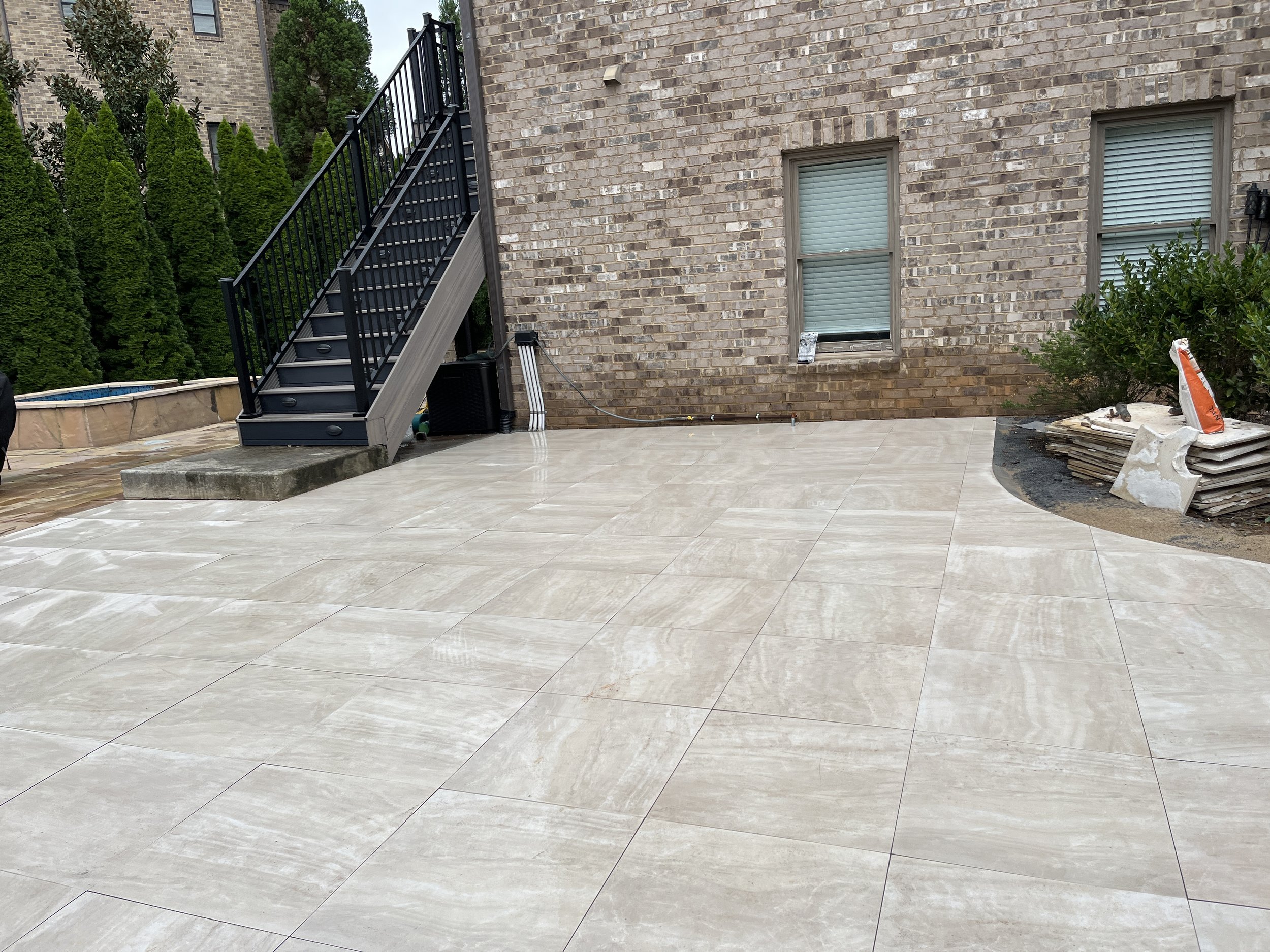 Backyard patio area with large beige tiles, a brick house wall with two windows, a staircase leading to an upper level, and outdoor construction materials on the ground.