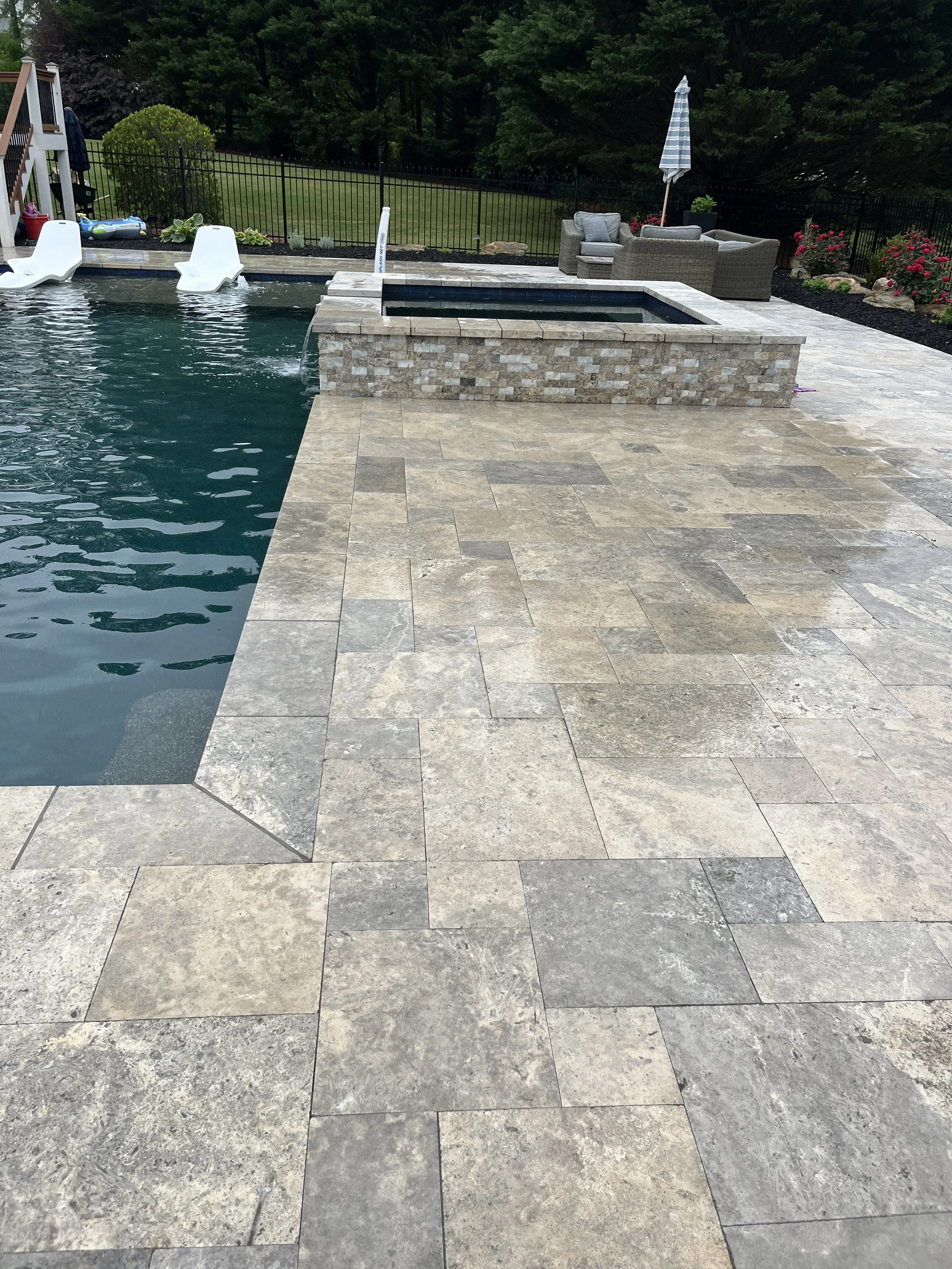 A backyard pool area with beige stone tiles, white pool floats, and a hot tub built with beige bricks. There are garden chairs with cushions and a large umbrella on the patio. A black fence surrounds the yard with trees and plants in the background.