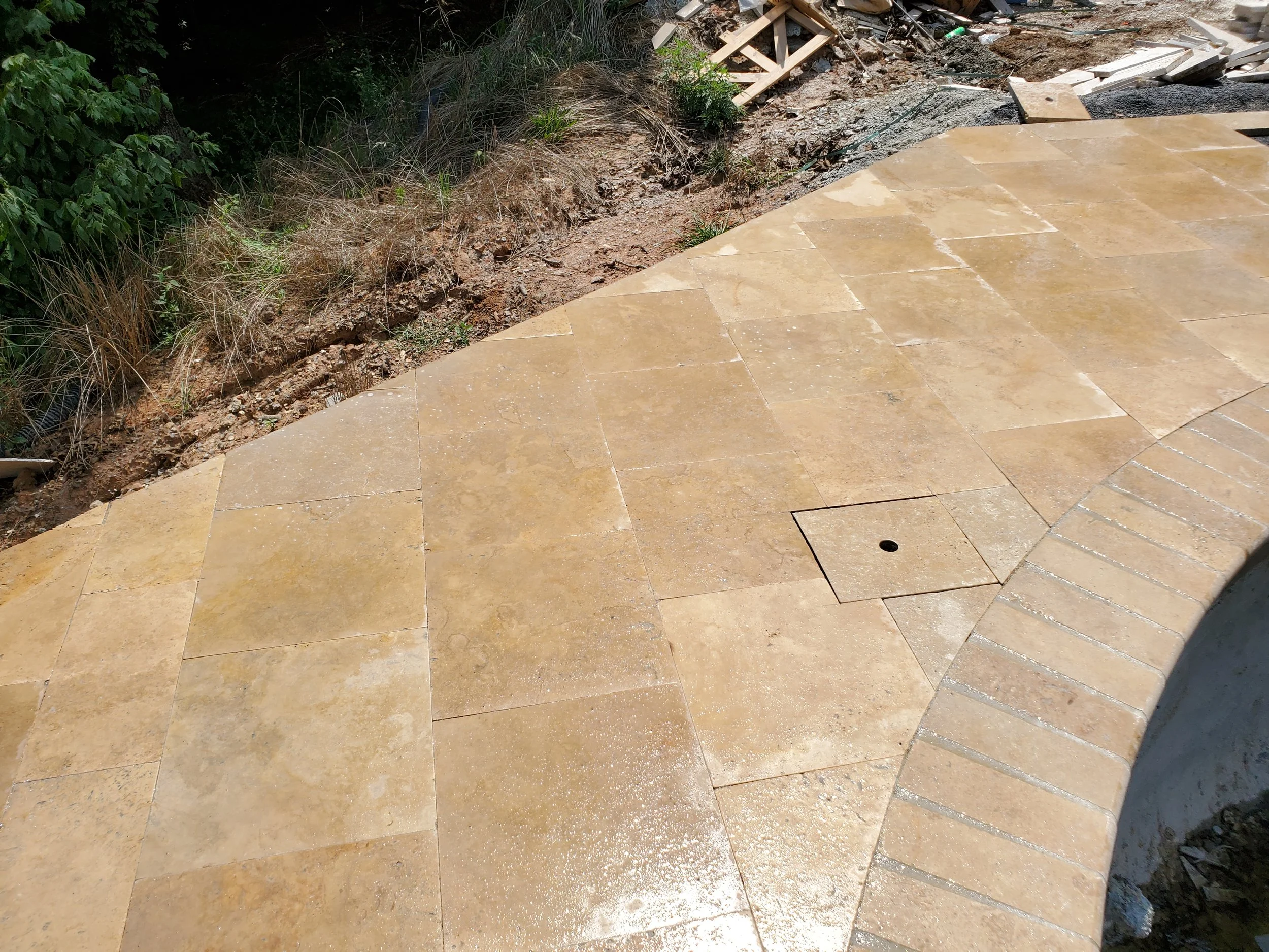 Part of a tiled patio with some dirt and plants along the edge, and construction debris including wood and gravel nearby.
