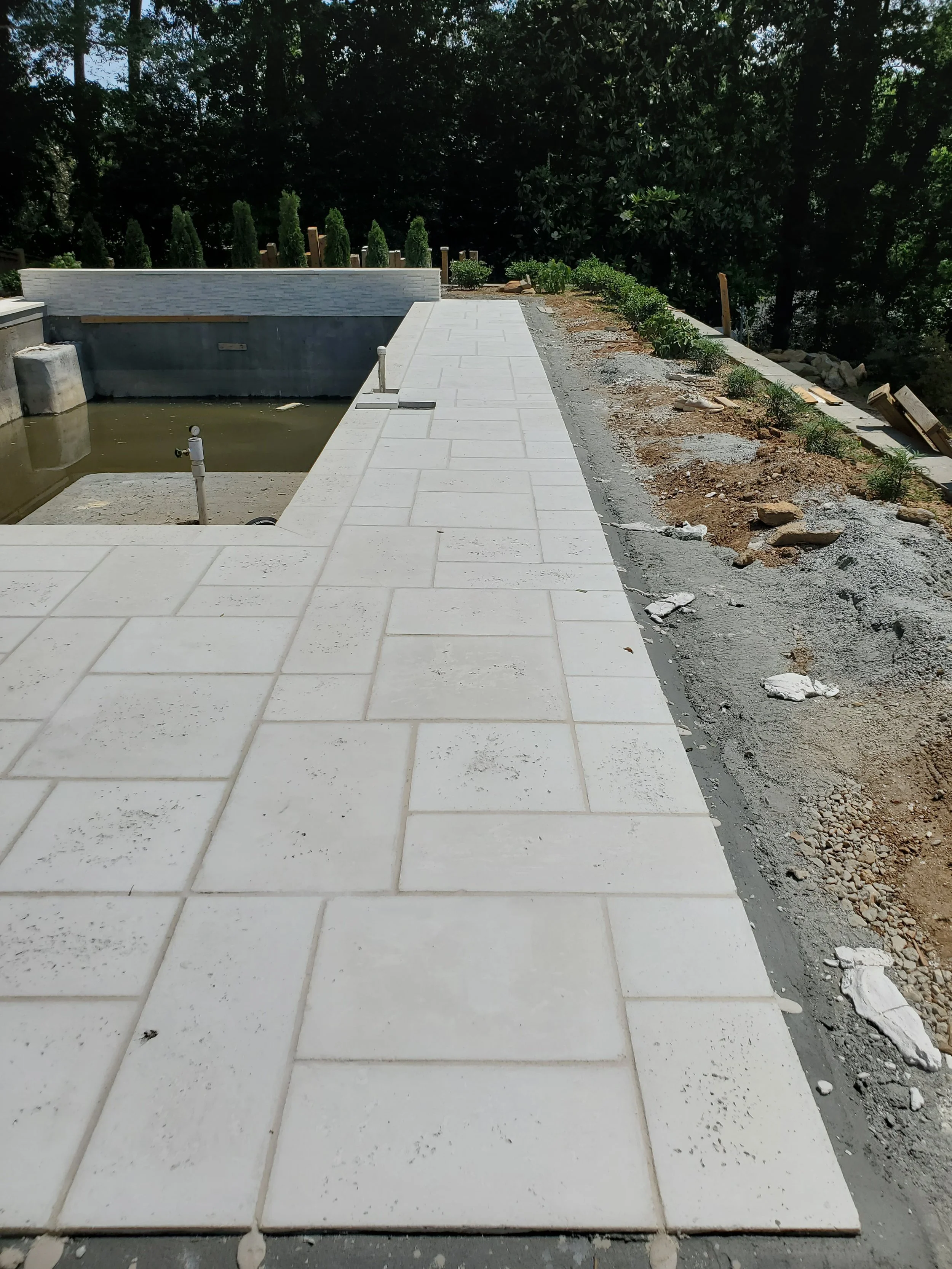 Construction site with newly installed white tiles on a walkway next to a partially built pool, dirt and construction debris on the ground, and lush green trees in the background.