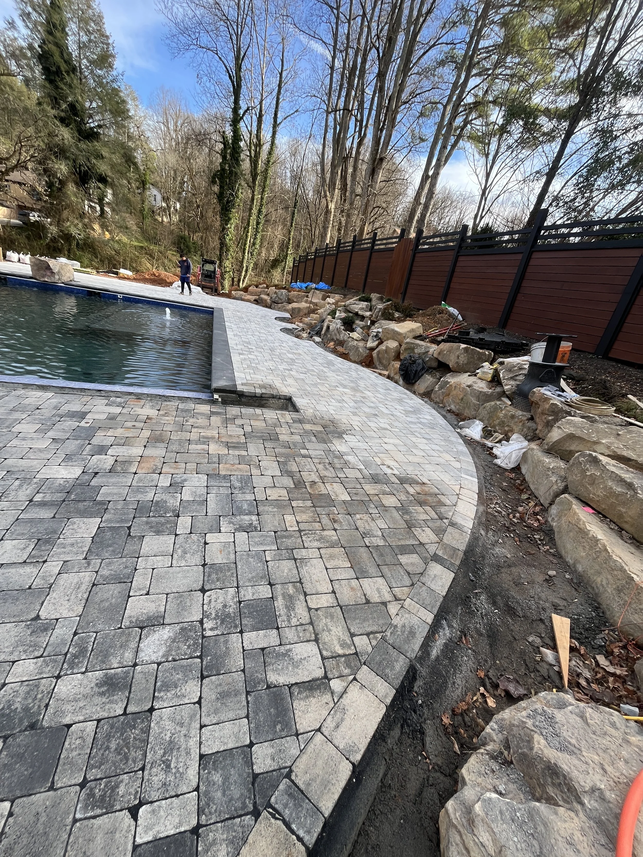 Construction work on a backyard patio with paving stones, rocks along the edge, and workers present. The scene includes a new stone walkway, a pool, and a wooden fence with trees in the background.