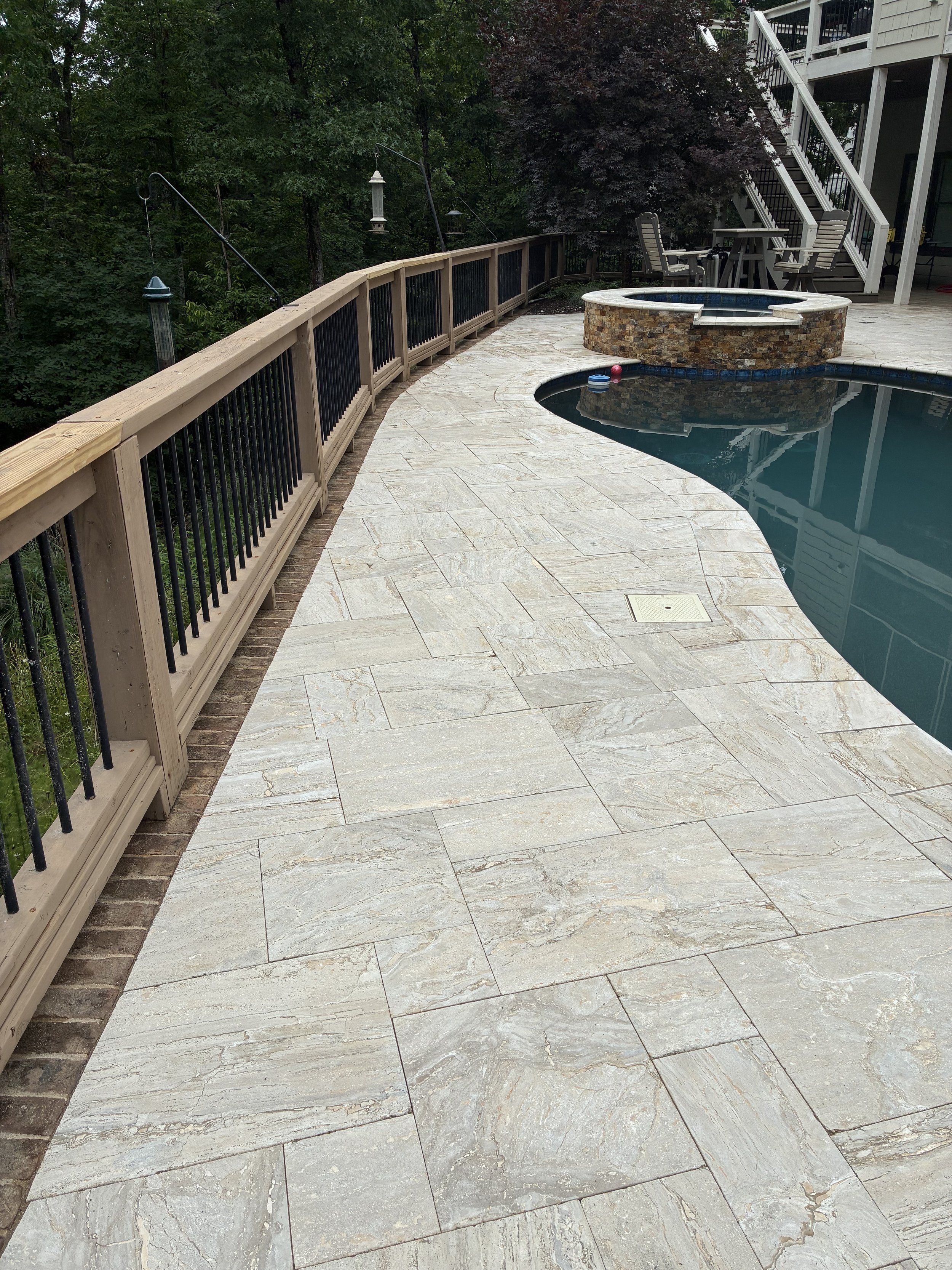 A backyard patio with a curved stone and tile swimming pool, a raised spa, outdoor seating, and a deck staircase, surrounded by trees and shrubs.