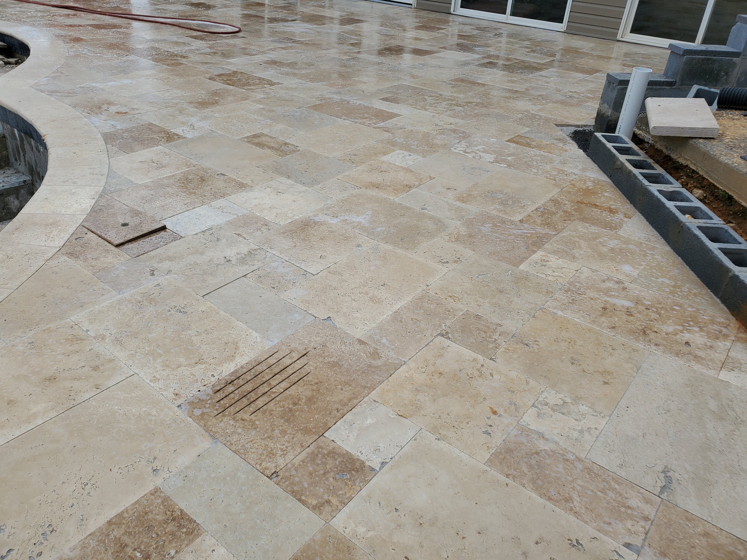Newly installed beige travertine stone patio with a curved edge, partially completed with some construction materials and a drain grate visible.