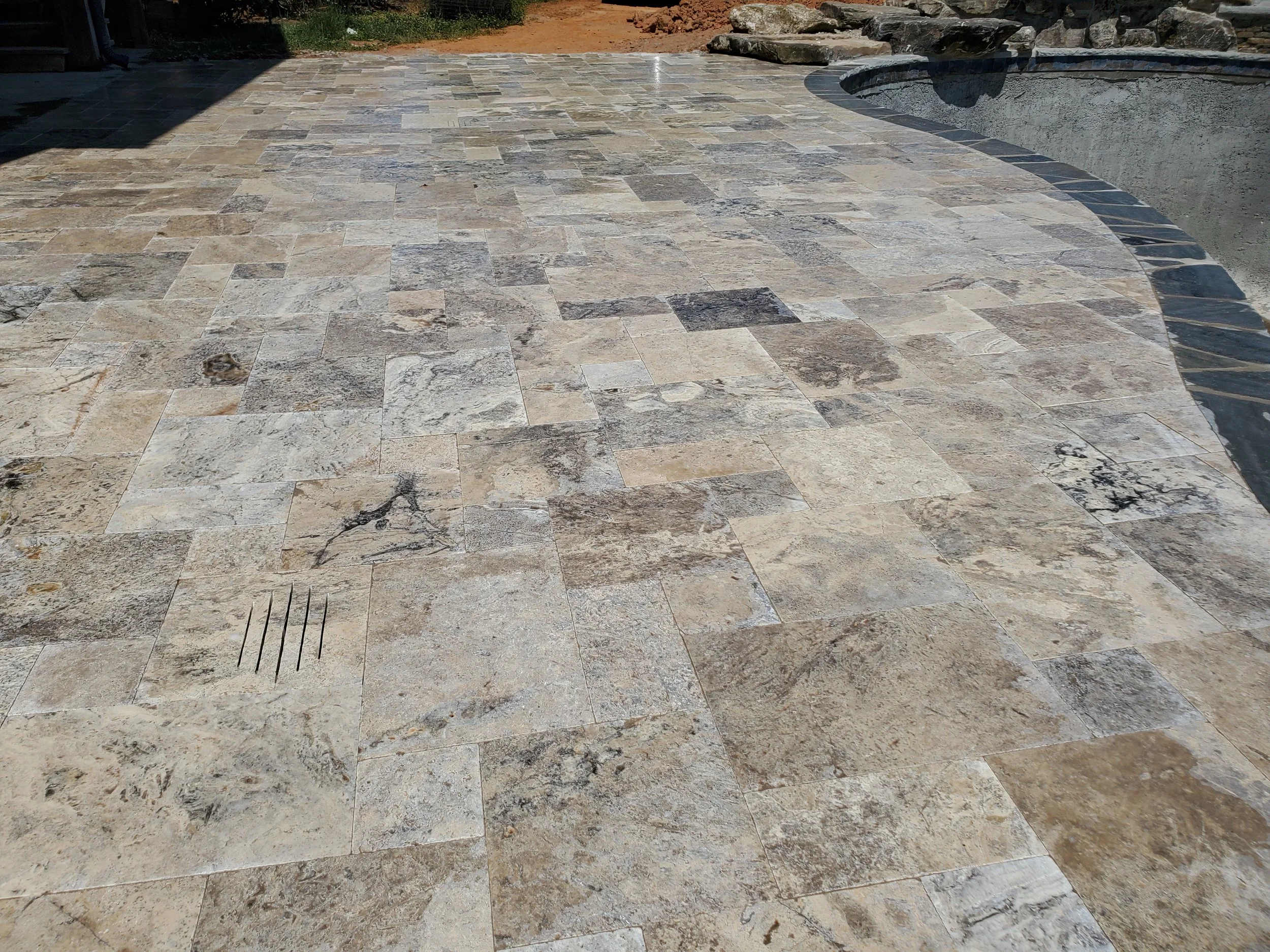 A newly constructed outdoor patio with beige and gray stone tiles, a pool edge on the right, a shadow casting over part of the patio, and an area of dirt and rocks in the background.