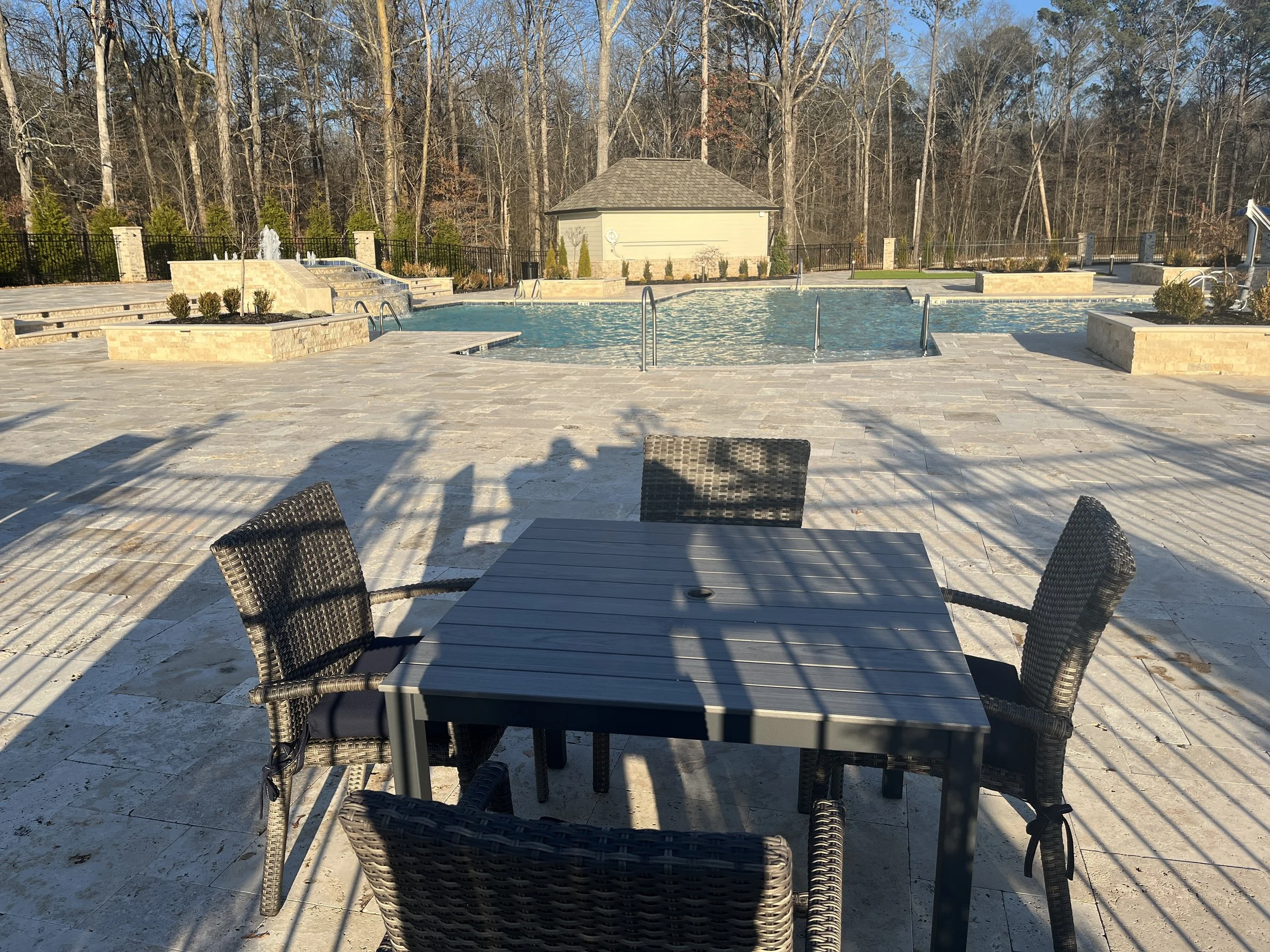 An outdoor swimming pool area with lounge chairs around the pool, a small building, black fence, trees in the background, and sunny weather.