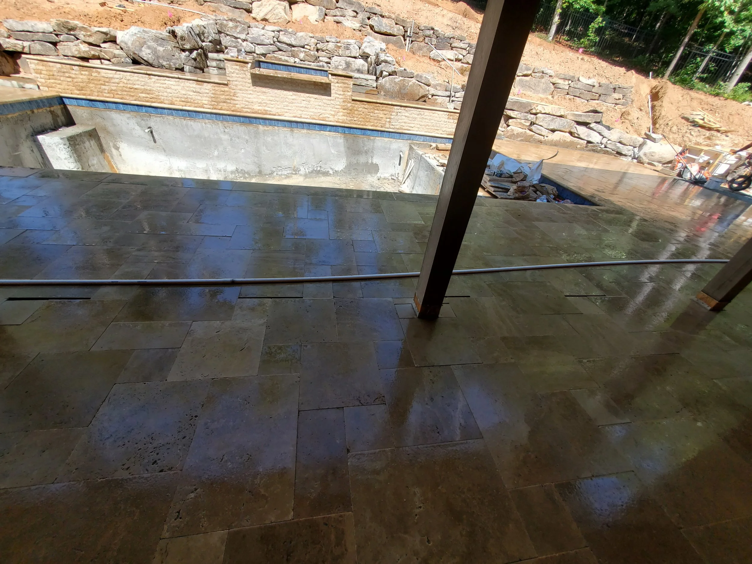 Construction site with a concrete pool, stone retaining walls, and unfinished landscaping, viewed from indoors through a sliding glass door.