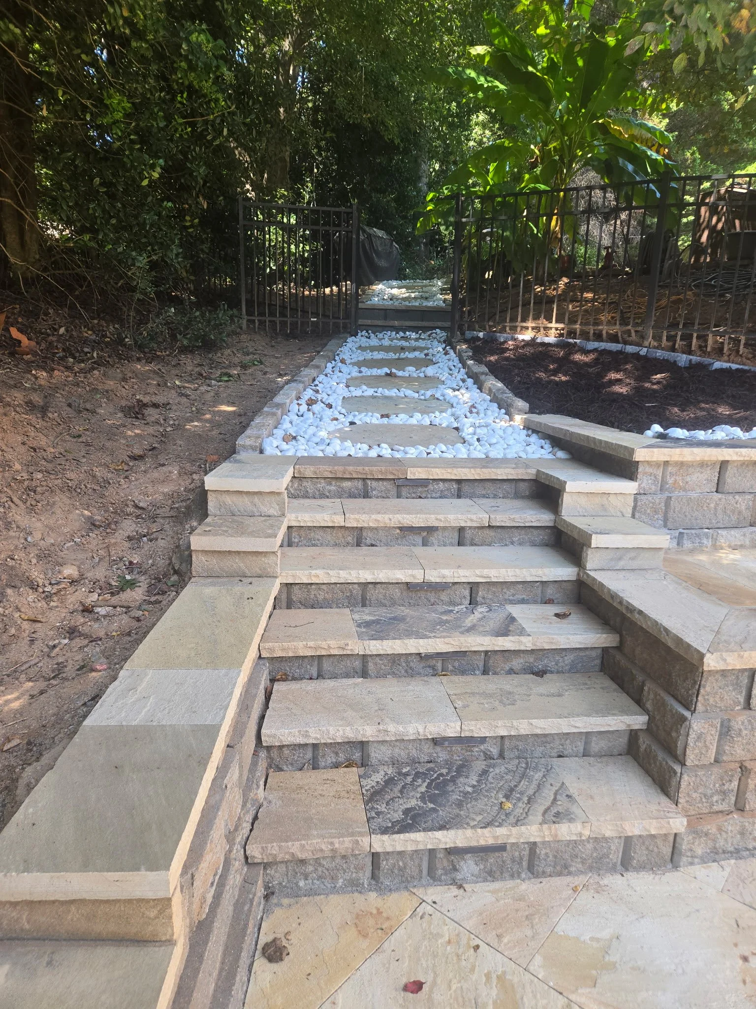 Newly constructed stone steps leading up to a small garden with a pathway filled with white decorative rocks, black metal fence, and lush green trees and plants in the background.