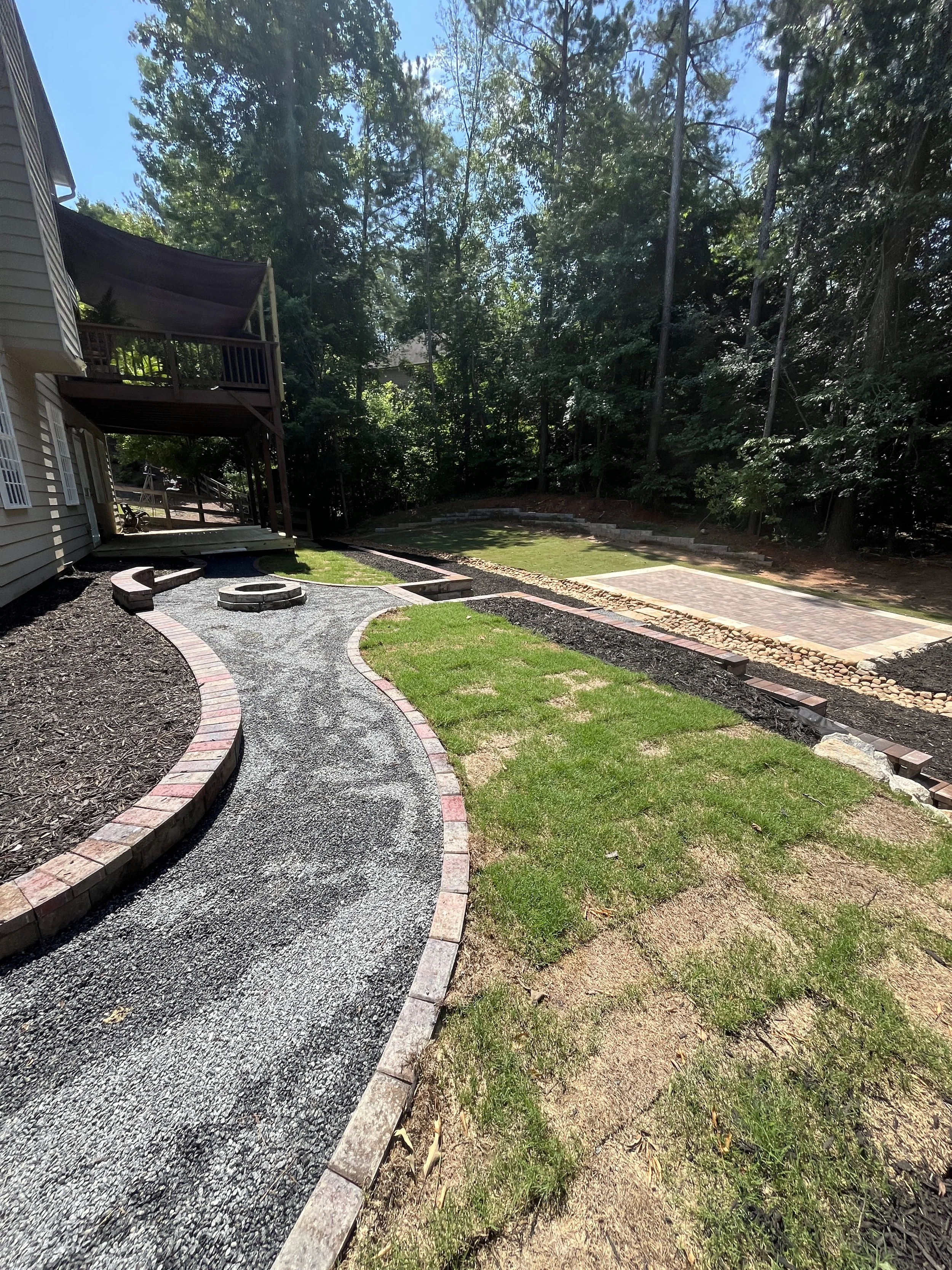 A backyard with a newly renovated winding gravel pathway edged with red bricks, a fire pit, and a lawn with small patches of grass. There is a raised deck attached to a house on the left and tall trees in the background.